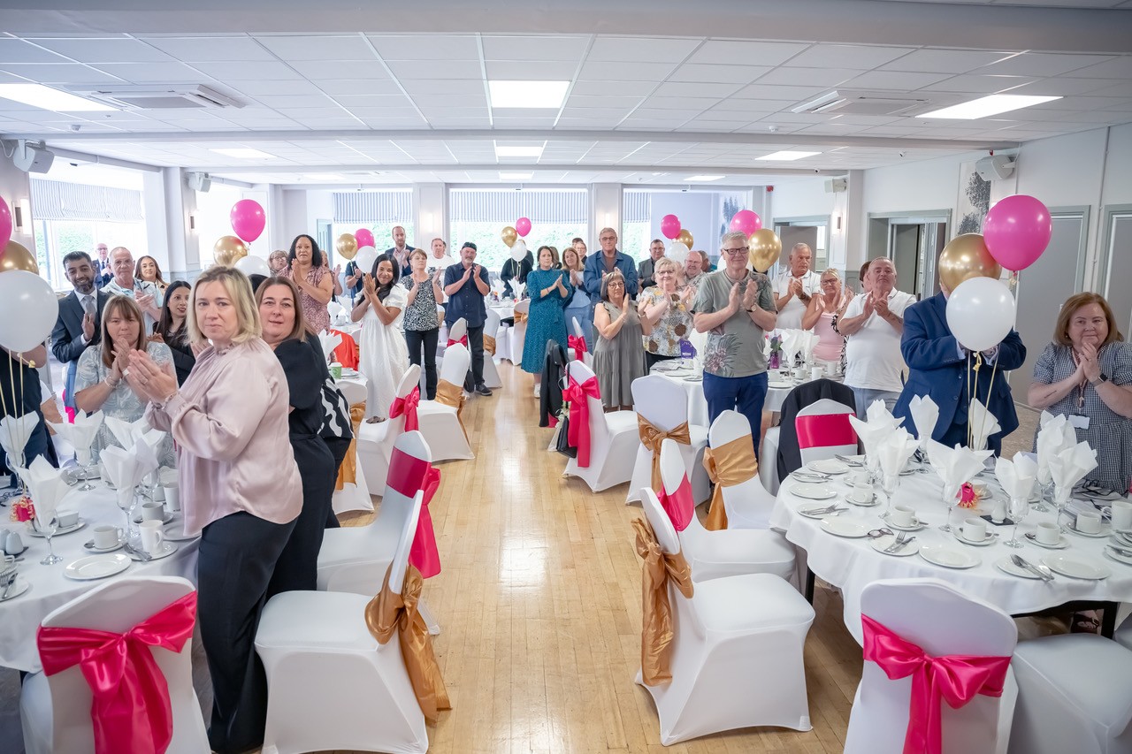 People stood behind tables lapping. There are pink, gold and white balloons on the tables