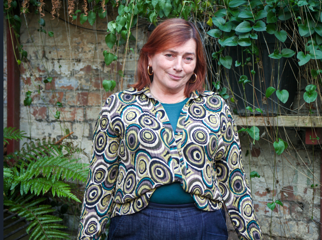 A person standing outdoors in front of a weathered brick wall covered with hanging vines and leafy plants. They are wearing a patterned button-up shirt with circular designs in green, yellow, and black tones over a teal top, paired with dark trousers. Ferns and trailing greenery frame the lower part of the scene.