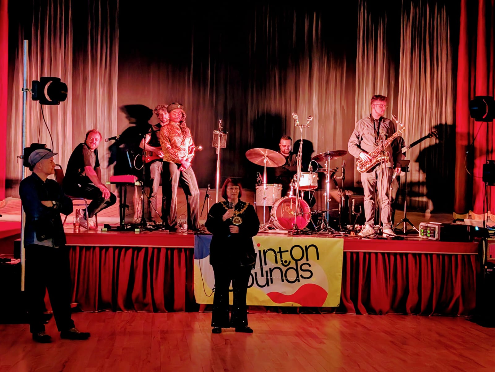 A live band performs on a stage in front of dark curtains under stage lighting. Musicians play instruments including drums, saxophone, and electric guitar. A woman with a mayoral chain stands on the floor in front of the stage, addressing the audience. A ‘Swinton Sounds’ banner hangs on the stage front.
