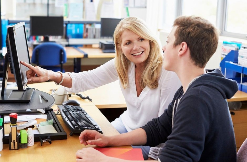 A woman smiles and points to a computer screen. She is sat next to a young man wearing a navy blue hoody, who is looking at the screen