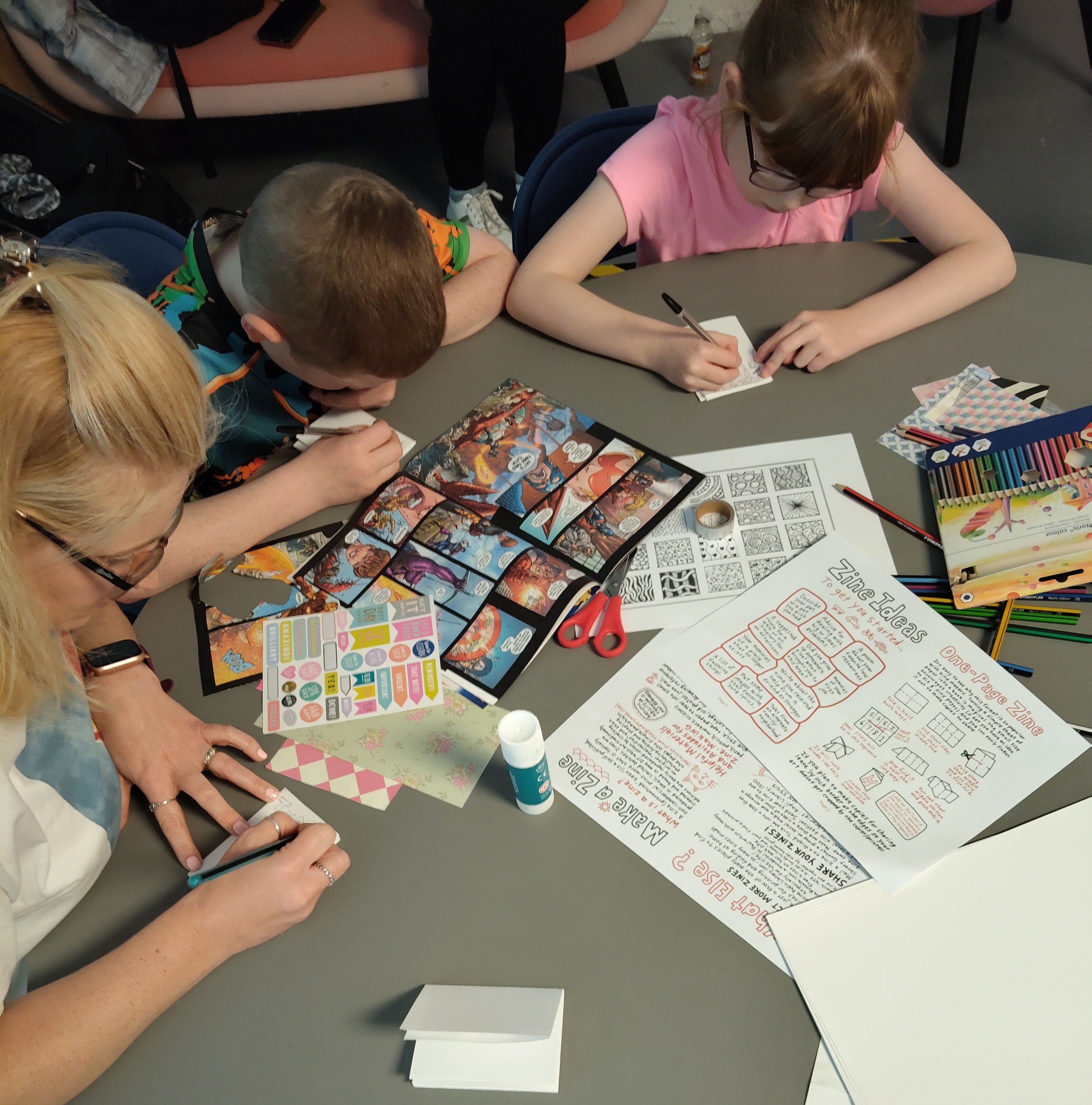 Two children and an adult sit around a circular table engaged in a zine-making workshop. The table is scattered with colourful craft supplies, including comic book pages, stickers, pencils, and instruction sheets. The children are focused on drawing and cutting paper, while the adult assists.