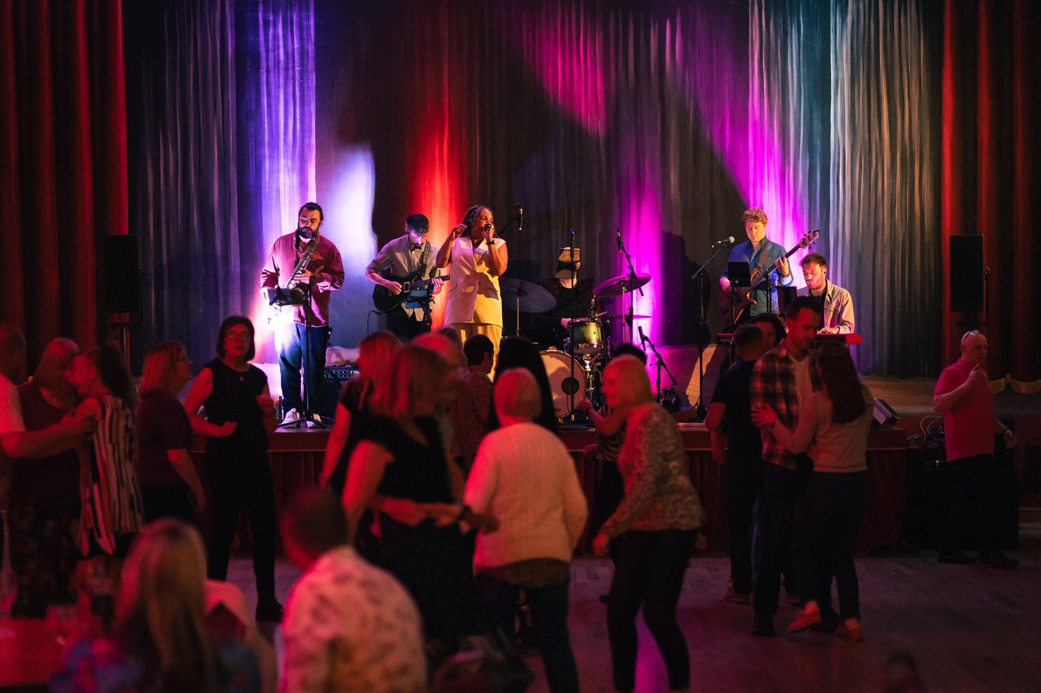 A group of people performing on a stage, with a theater curtain in the background. People in the foreground are dancing to the band.