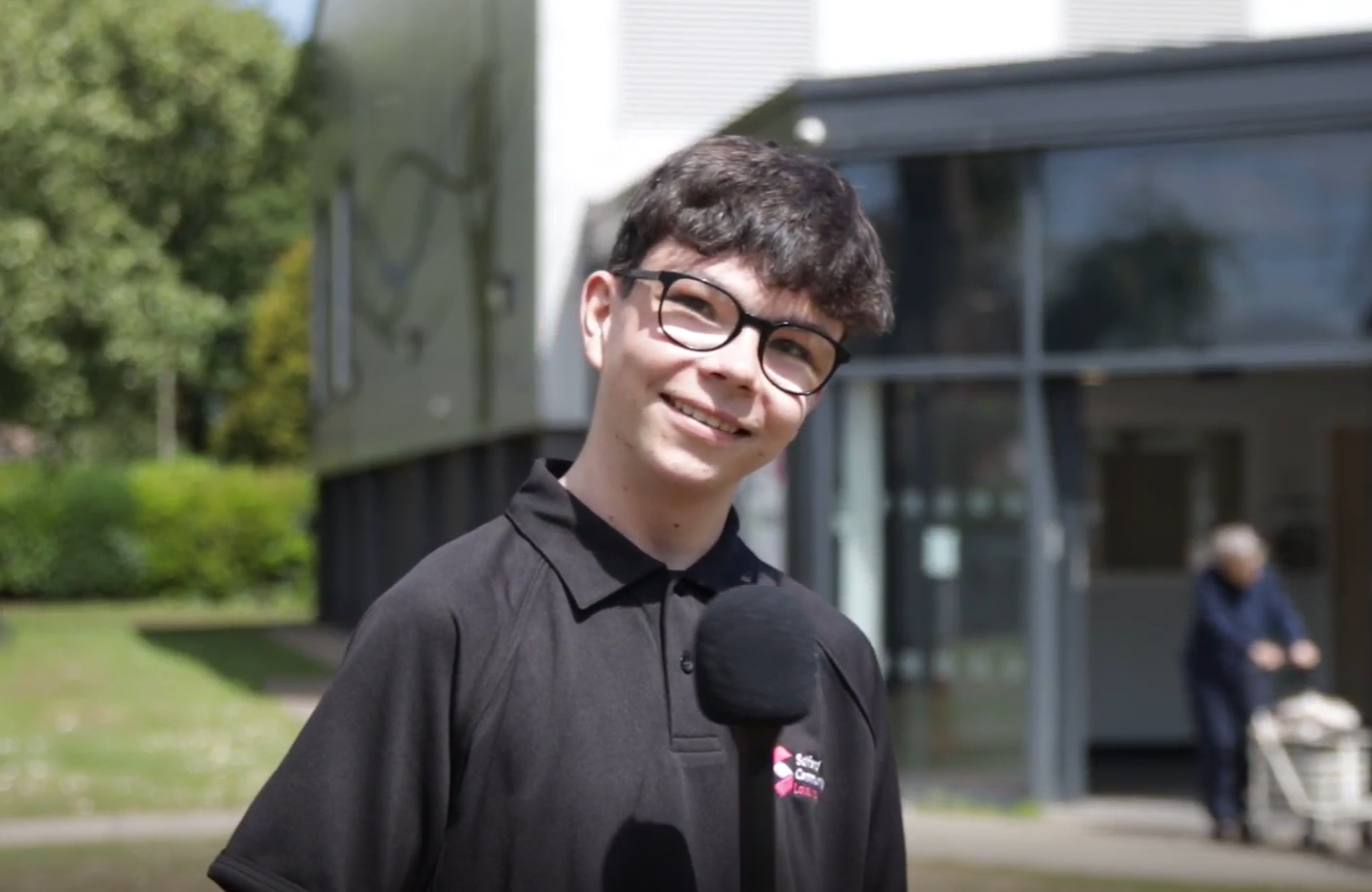 A young man wearing glasses and a black polio shirt stands smiling in front of a building and trees