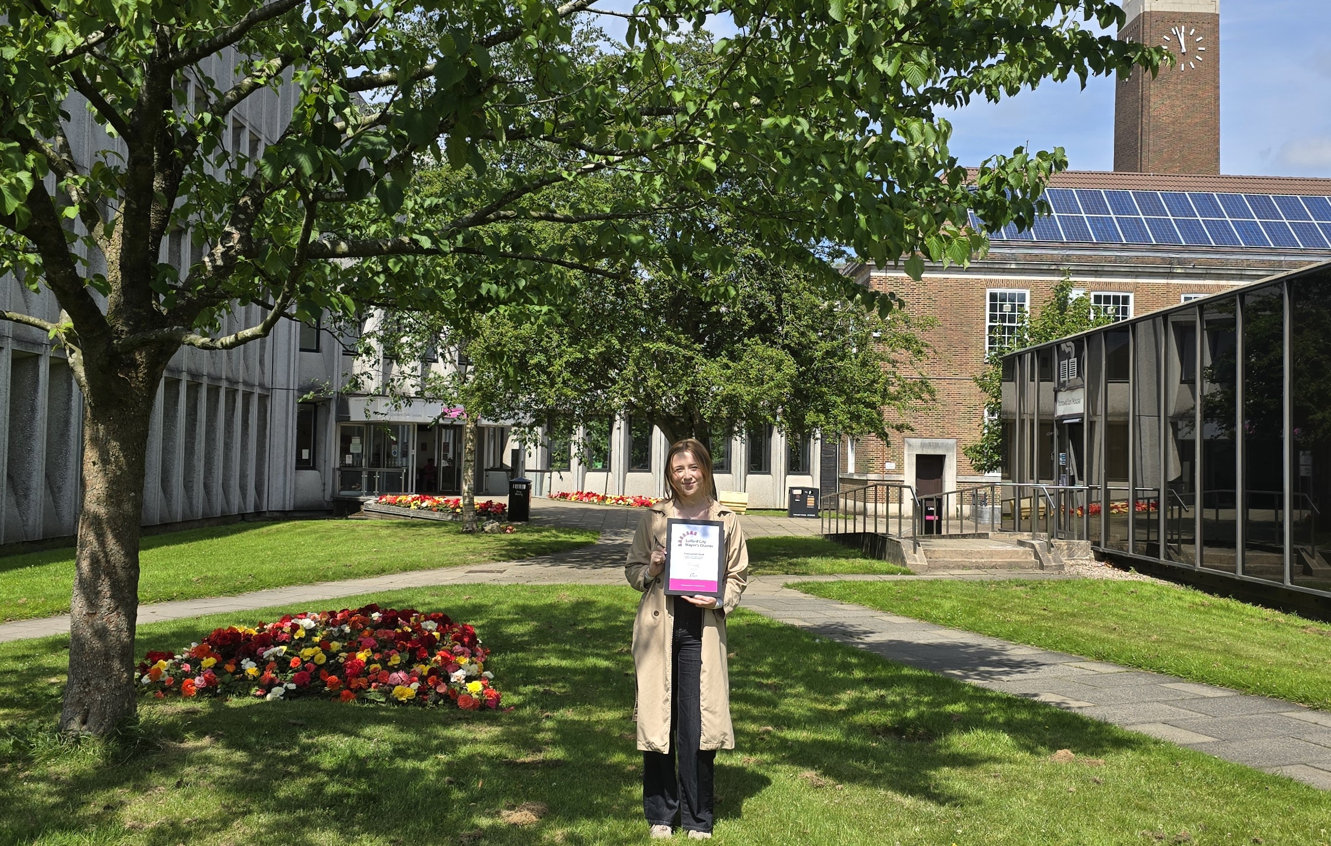 A woman in a brown coat and black trousers standing smiling and holding a framed certificate. She is stood outside on a grassed area. A tree is visible in the foreground, with buildings and a clock tower in the background.