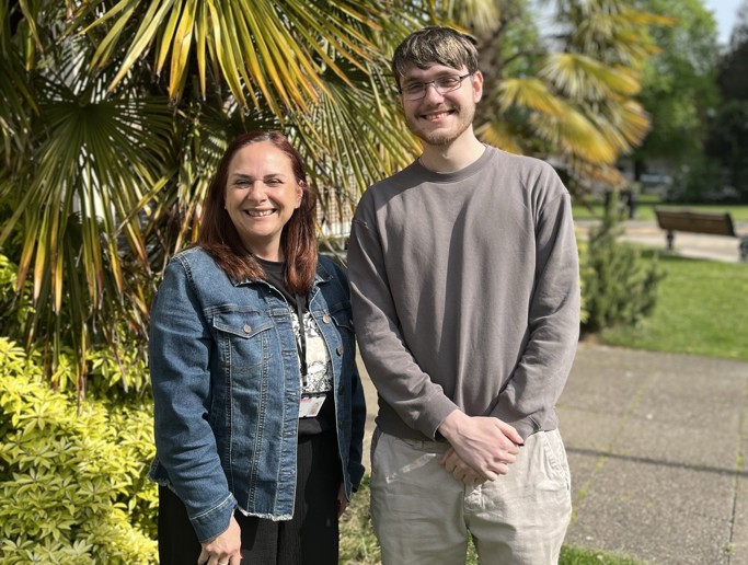 A woman and a man stand outside in front of some trees, smiling.