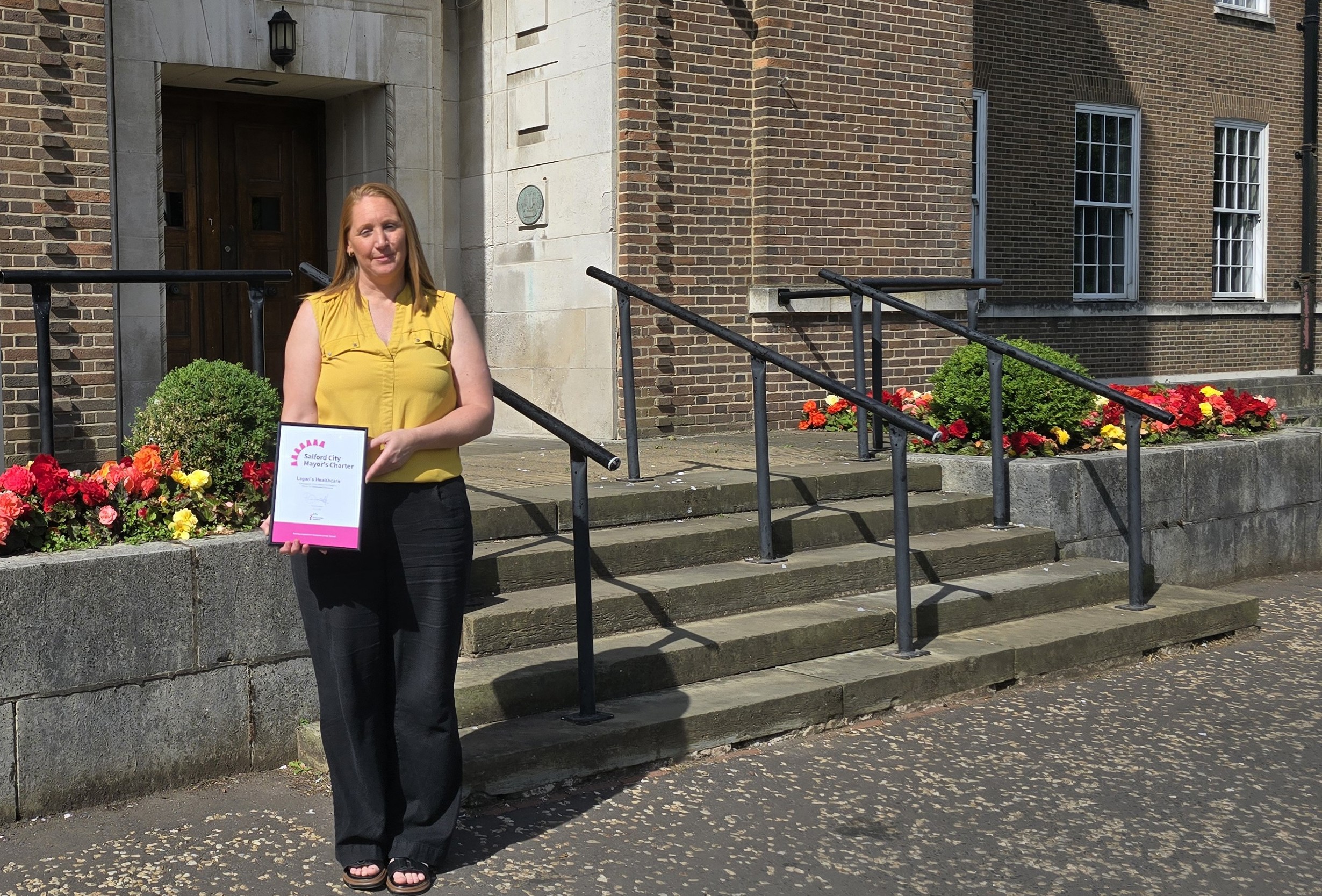 A lady stands holding a framed certificate, smiling. She is wearing a yellow vest top and black trousers, and stood in front of some stone steps, railings, flowers and shrubbery.