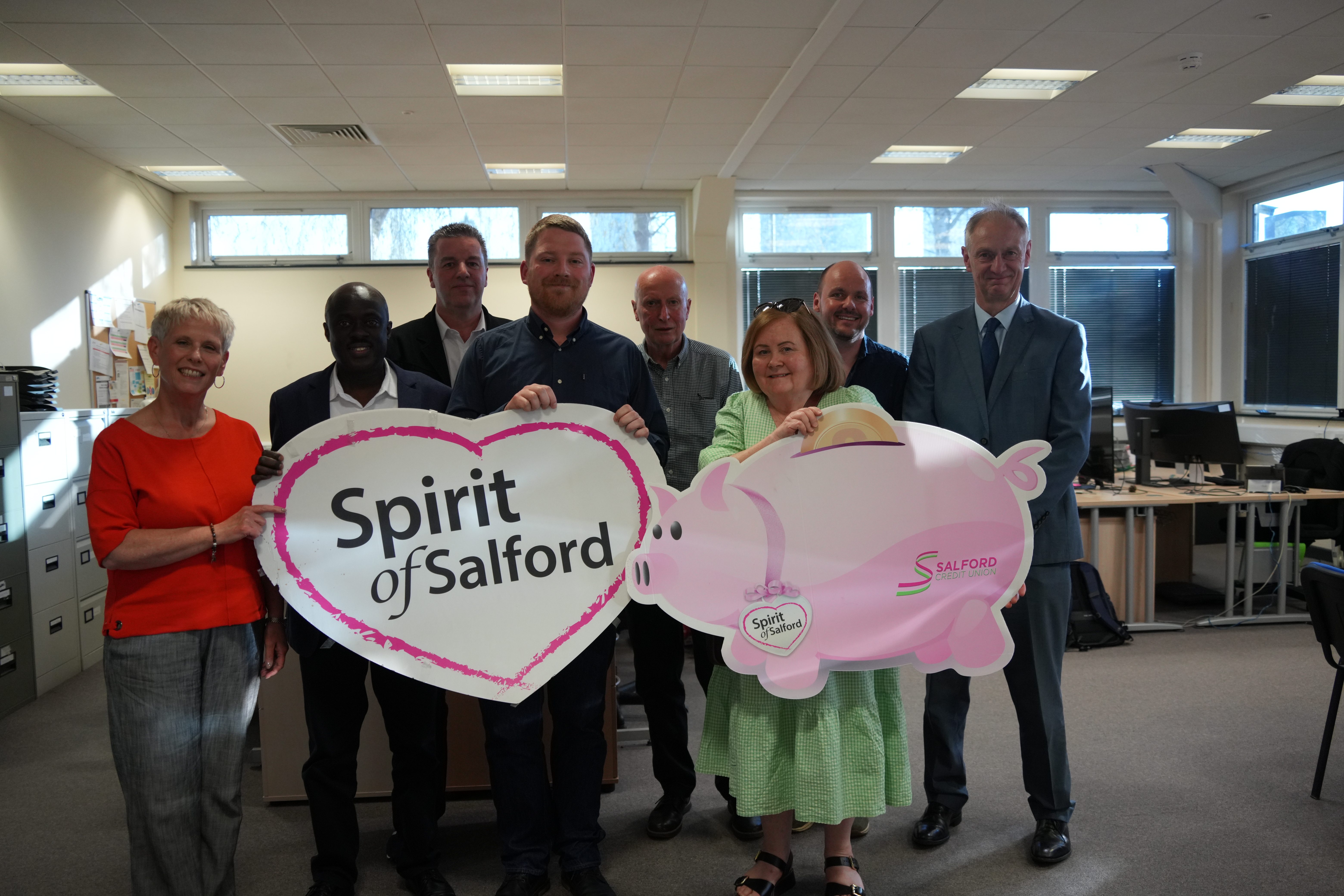 A group of people stood with a cardboard piggy bank and a heart which says Spirit of Salford
