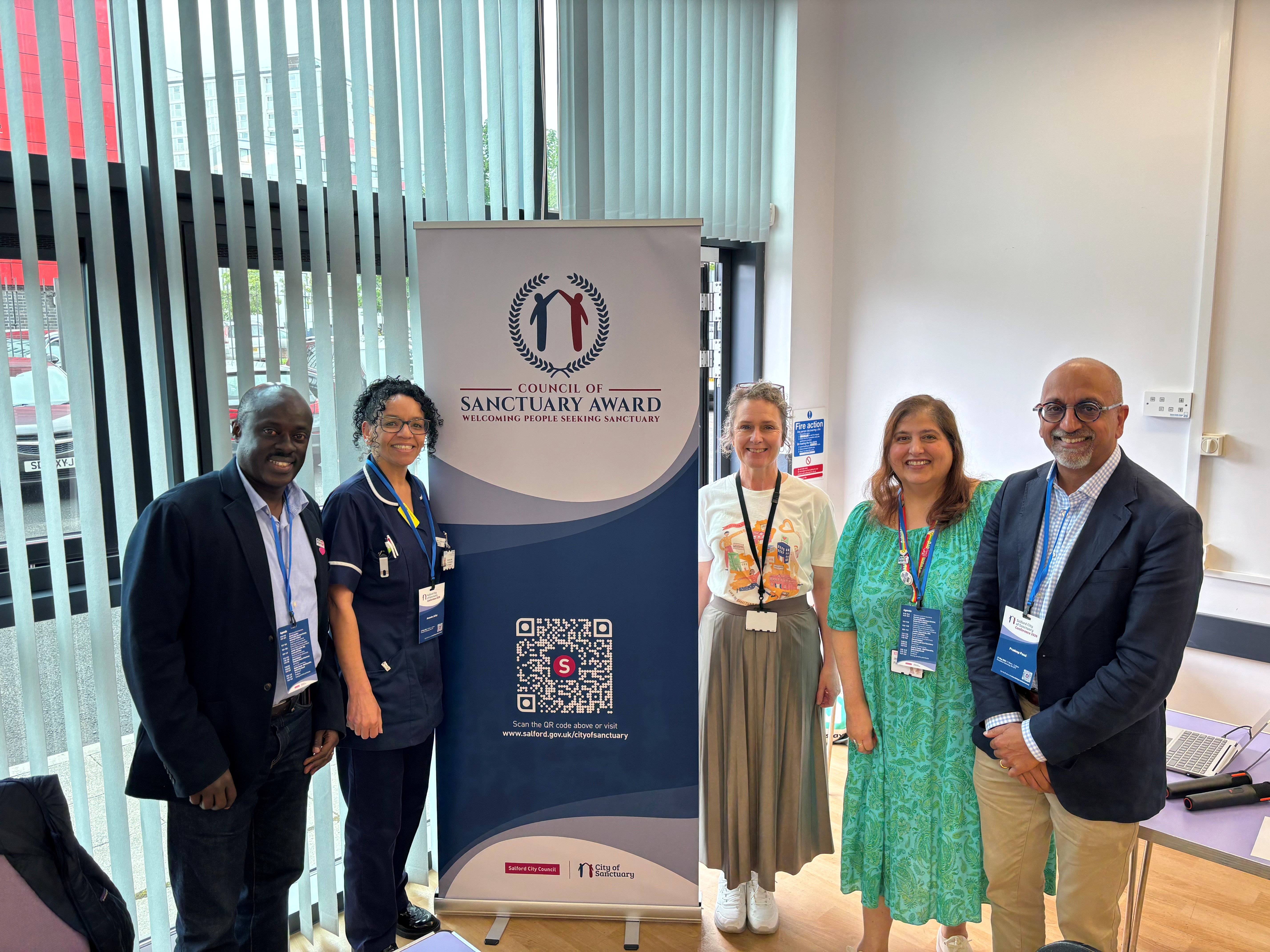 A group of people are stood next to a banner to recognise Salford receiving official accreditation as City of Sanctuary