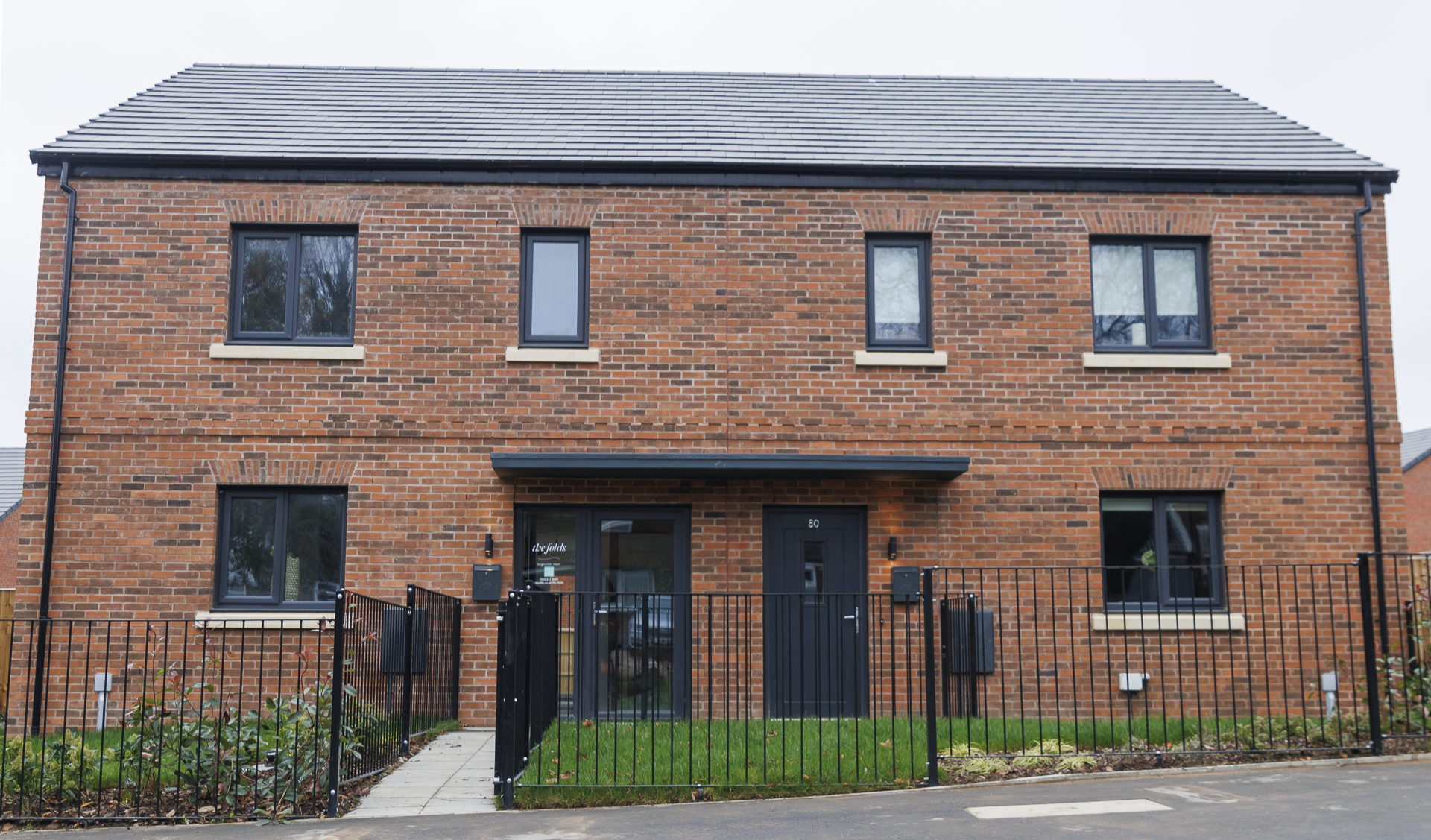 A completed modern red-brick semi-detached house with black-framed windows and doors, set behind a neat black metal fence and landscaped front garden. The house has a clean, finished appearance, with a concrete path leading to two front doors under a shared canopy.