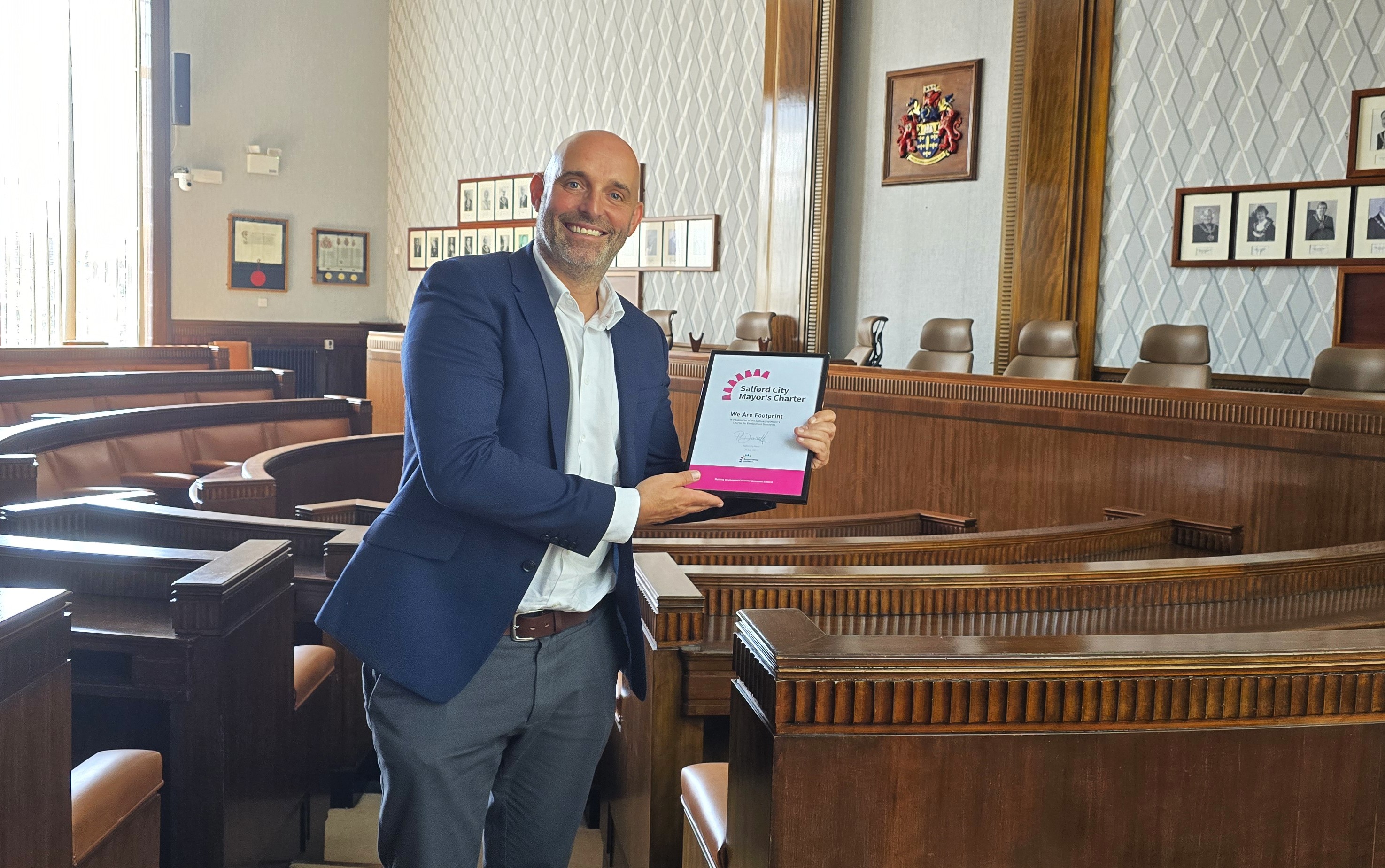 A man stands smiling and holding a framed certificate. He is wearing a navy jacket over a white dress shirt, tucked into grey trousers. In the background, wooden seats and picture frames can be seen against a patterned wall.