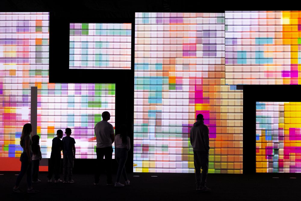 A group of silhouetted people, including adults and children, stand in front of large, brightly illuminated digital screens displaying colorful, abstract, pixel-like patterns in a dark room, resembling a modern immersive art installation.