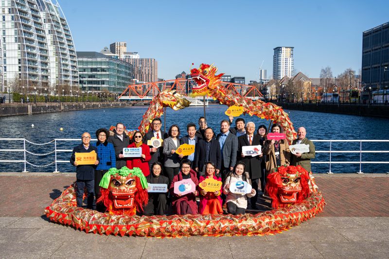 Partners for the UK Dragon Boat Festival stood together holding up signs from their organisation with red and gold dragons swirled around them. They are stood in front of the water at The Quays.
