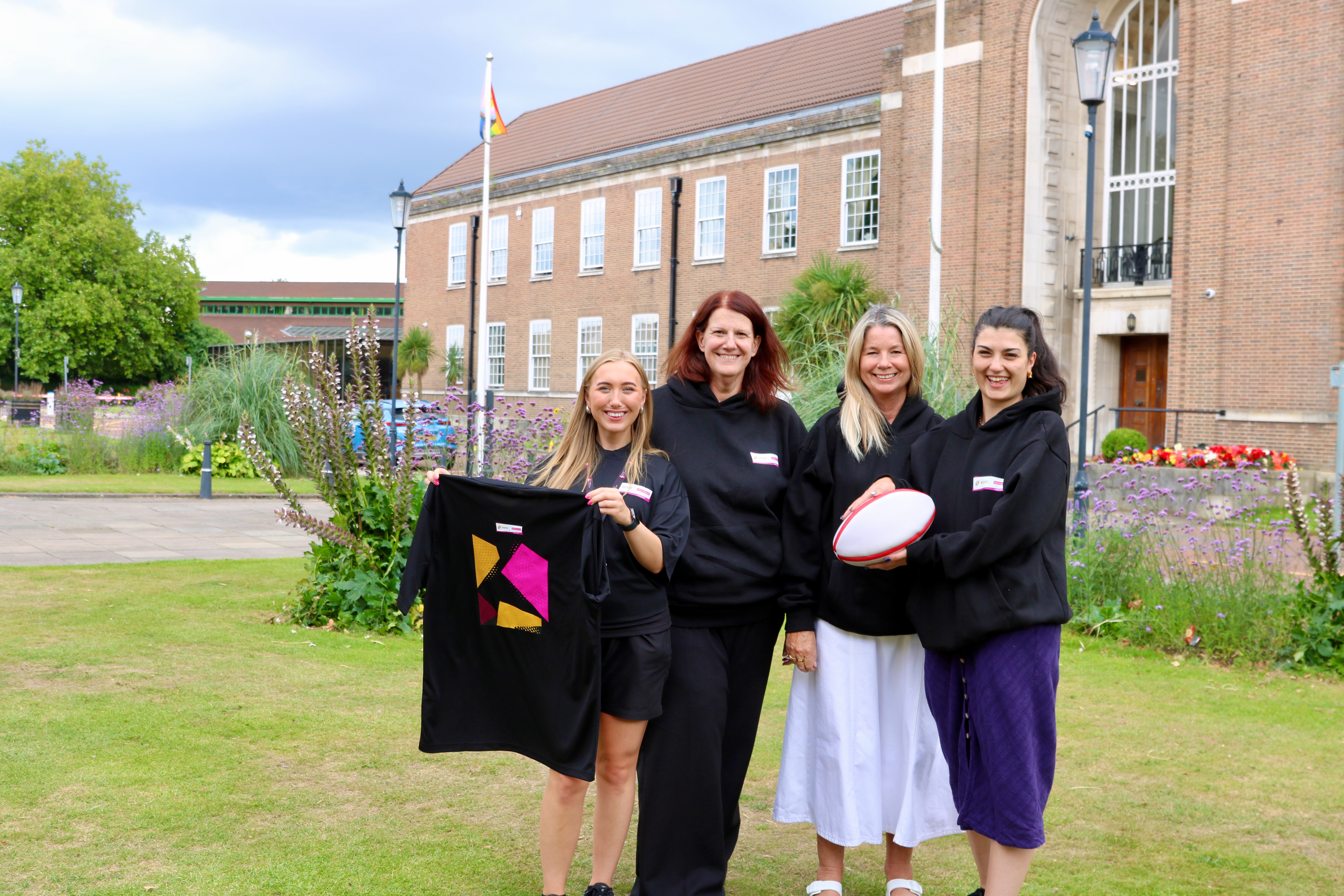 A group of 4 women smiling. One is holding a rugby ball and one is holding a t-shirt up