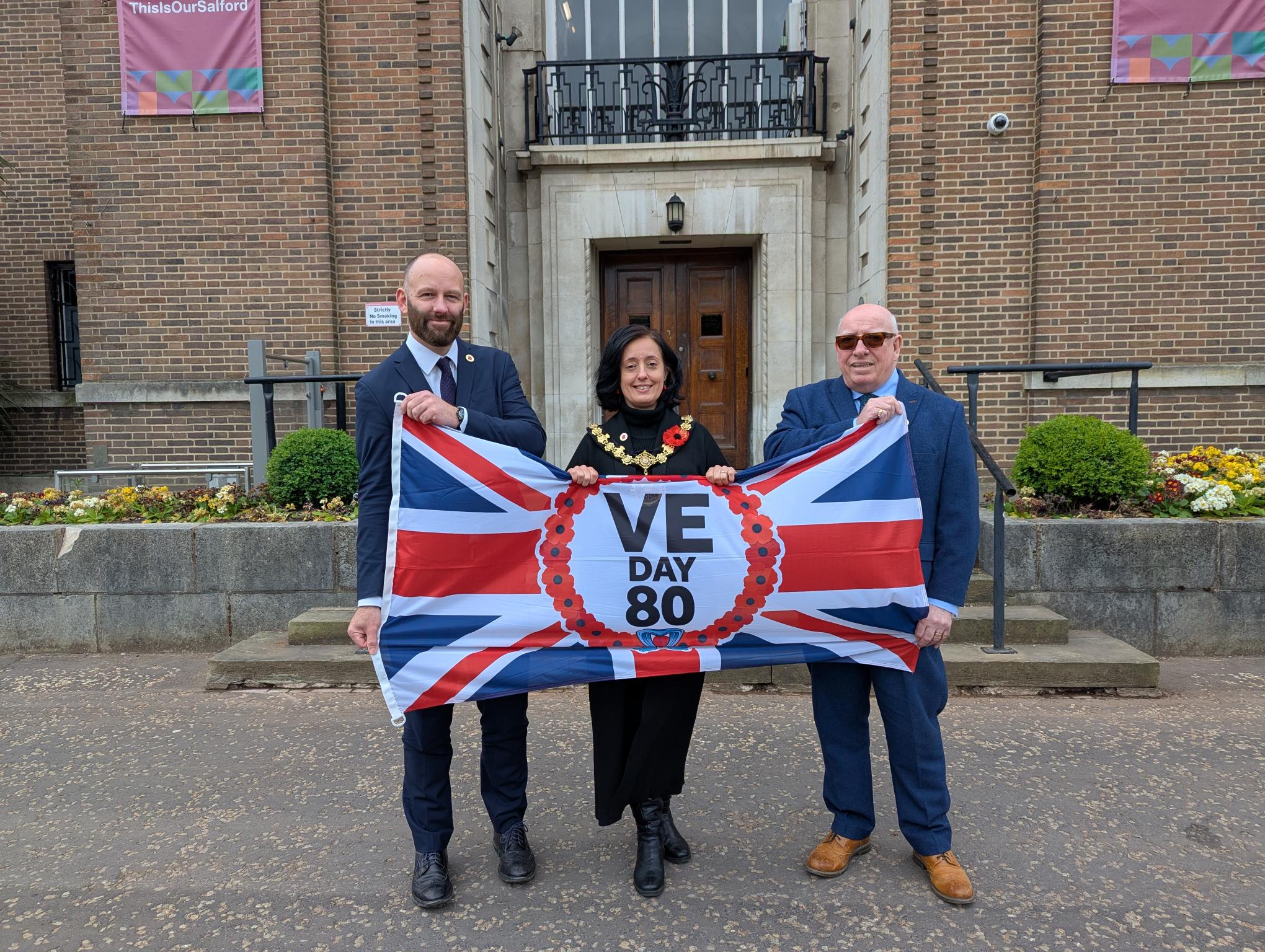 Salford City Mayor Paul Dennett, Salford Ceremonial Mayor Councillor Tanya Burch and Leader of the Opposition Councillor Robin Garrido holding VE Day 80 flag