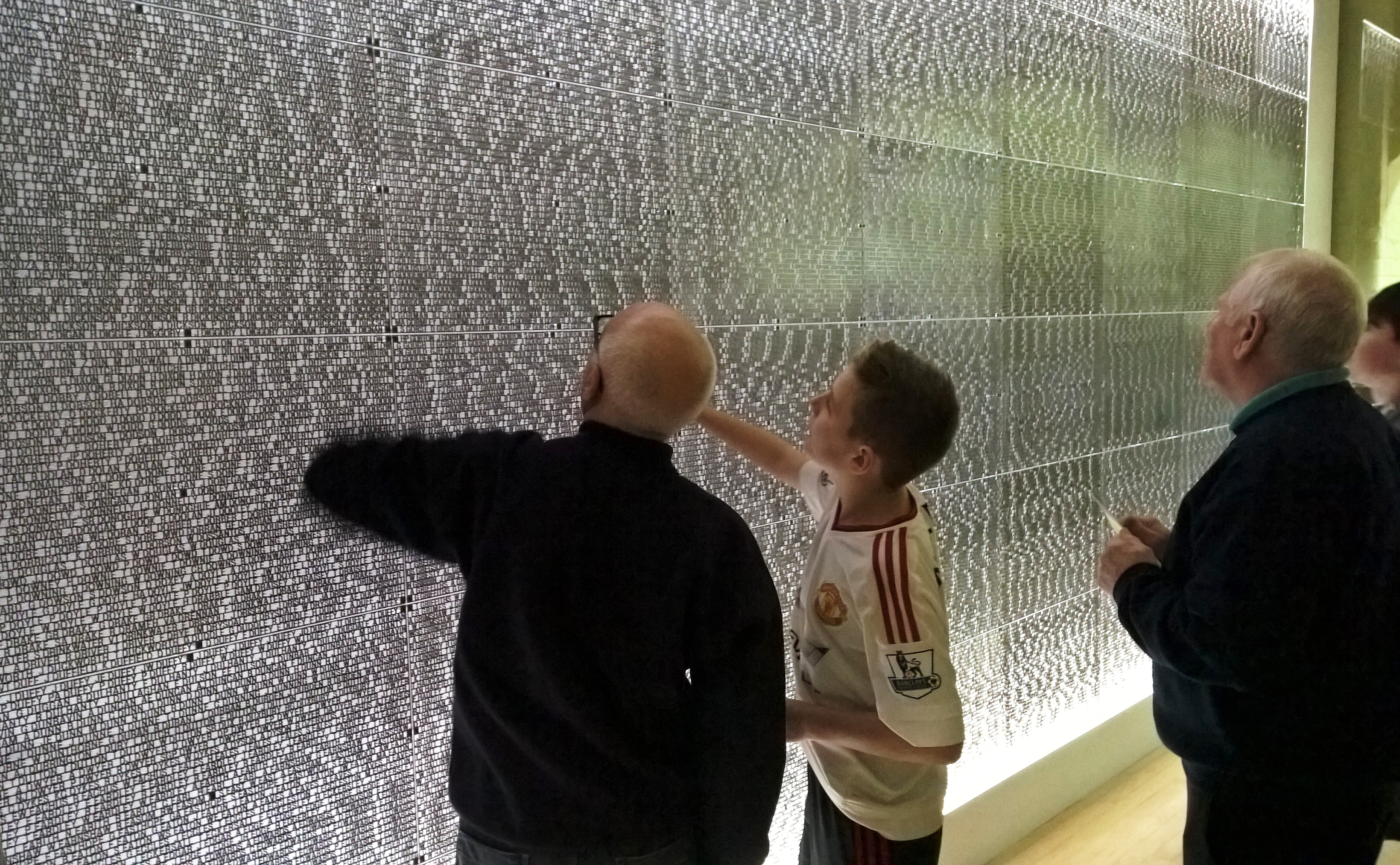 A group of people, including a boy in a football jersey and several men, examine and point at names engraved on the "Wall of Names" inside Salford Lads Club. The wall is covered with thousands of small, evenly spaced text entries, honouring past members of the historic club.