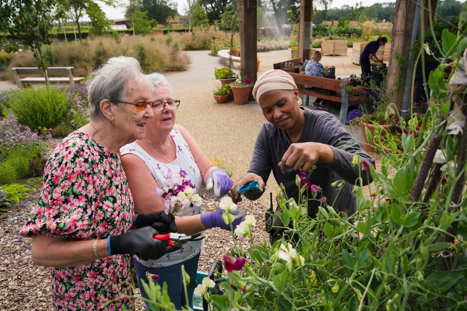 A group of women standing in RHS Bridgewater garden surrounded by plants and flowers, holding pruners looking at flowers. Plants and trees in the background.