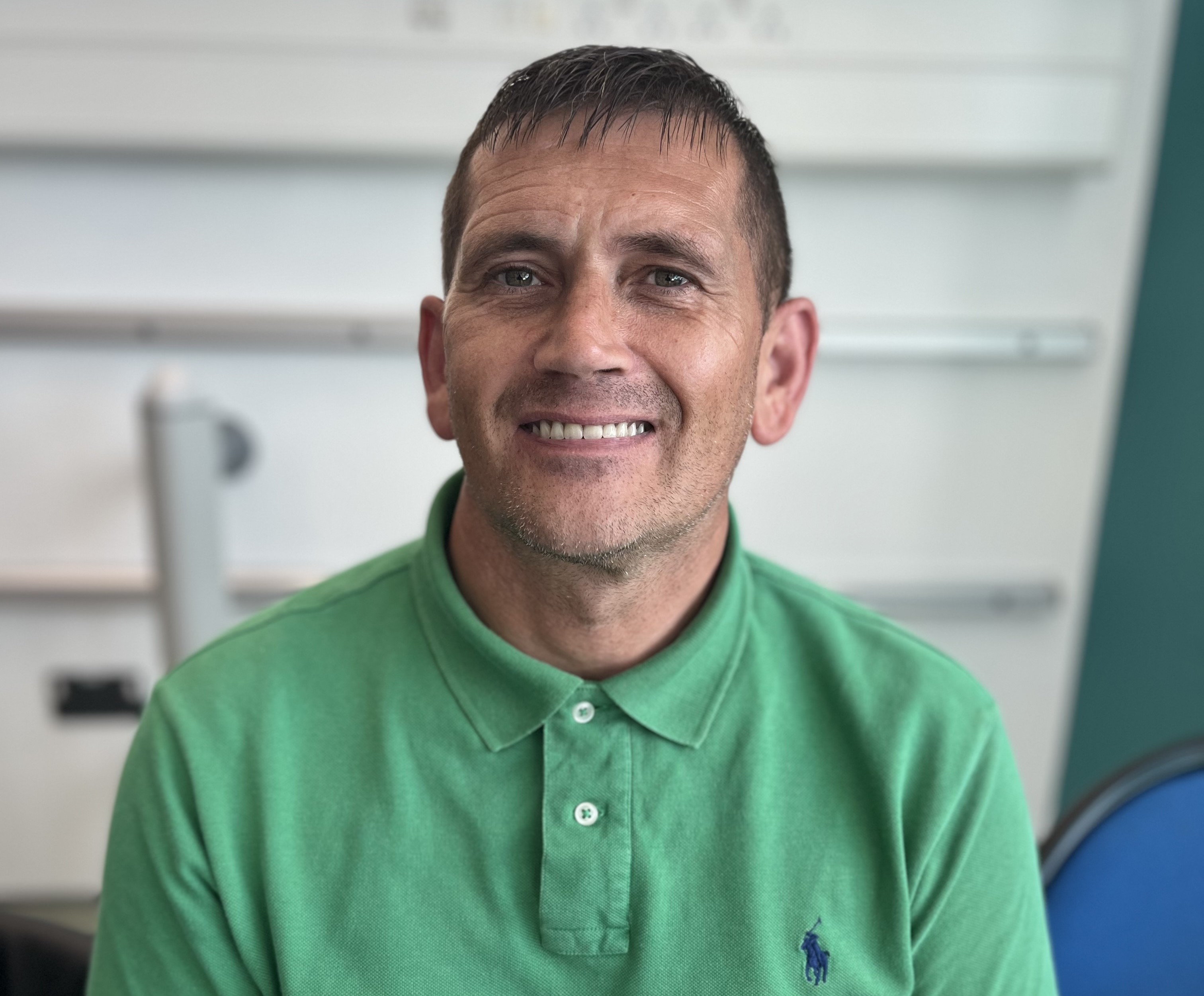 A man smiling in front of a blurred background. He is wearing a green polo shirt.
