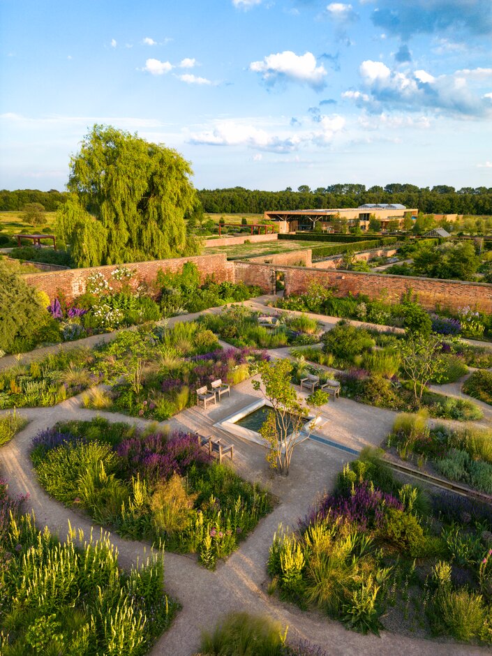 Aerial view of RHS Garden Bridgewater showing geometric flower beds, winding paths, benches and lush planting, with brick walls and woodland beyond.
