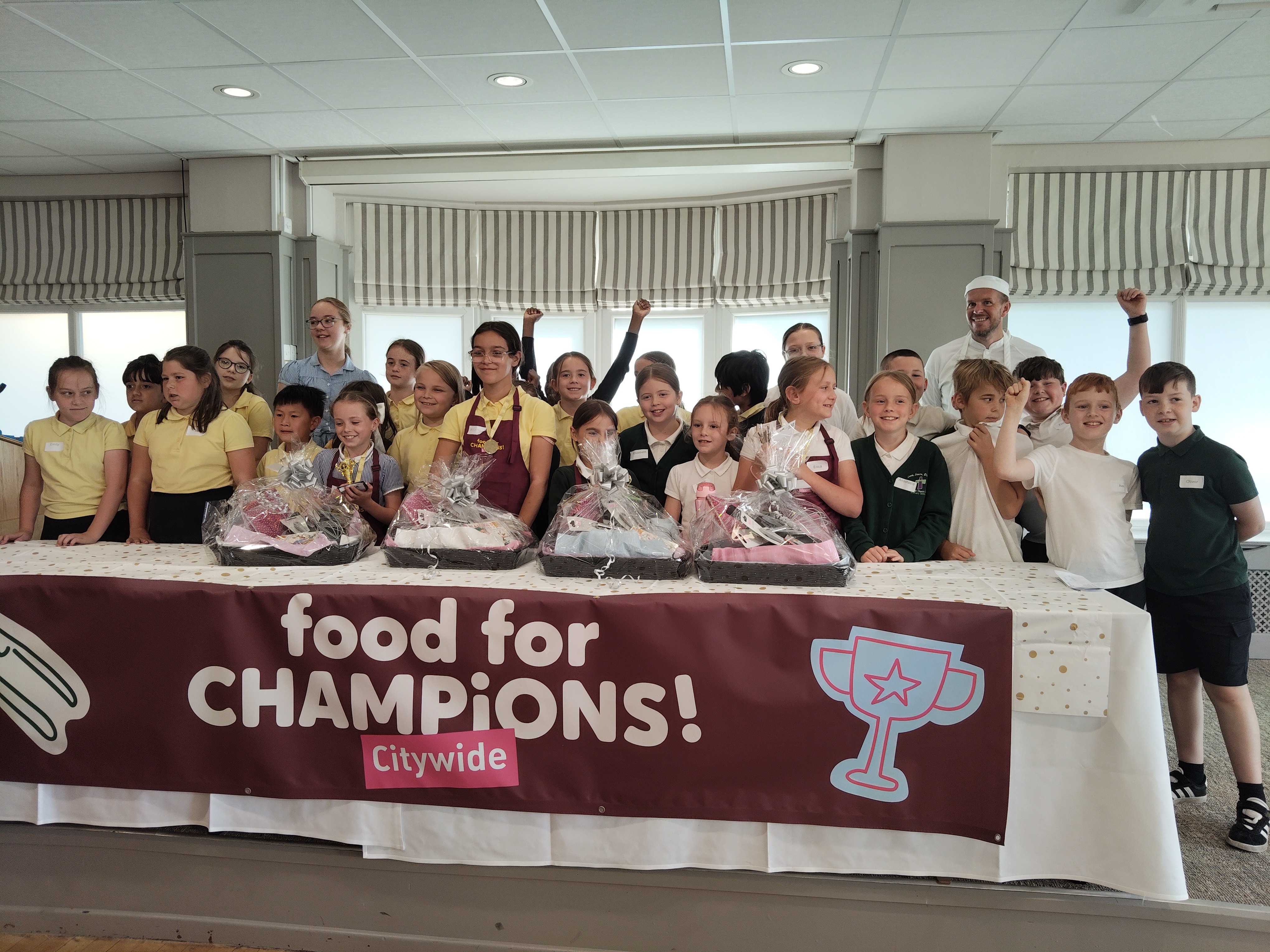 A large group of primary school children in uniform stand behind a table at Buile Hill Park Hall, smiling and cheering after a cookery competition. The table is decorated with a “Food for CHAMPiONS! Citywide” banner and holds prize hampers wrapped in cellophane. Two adults, including a chef in white uniform, stand at the back, celebrating with the children.