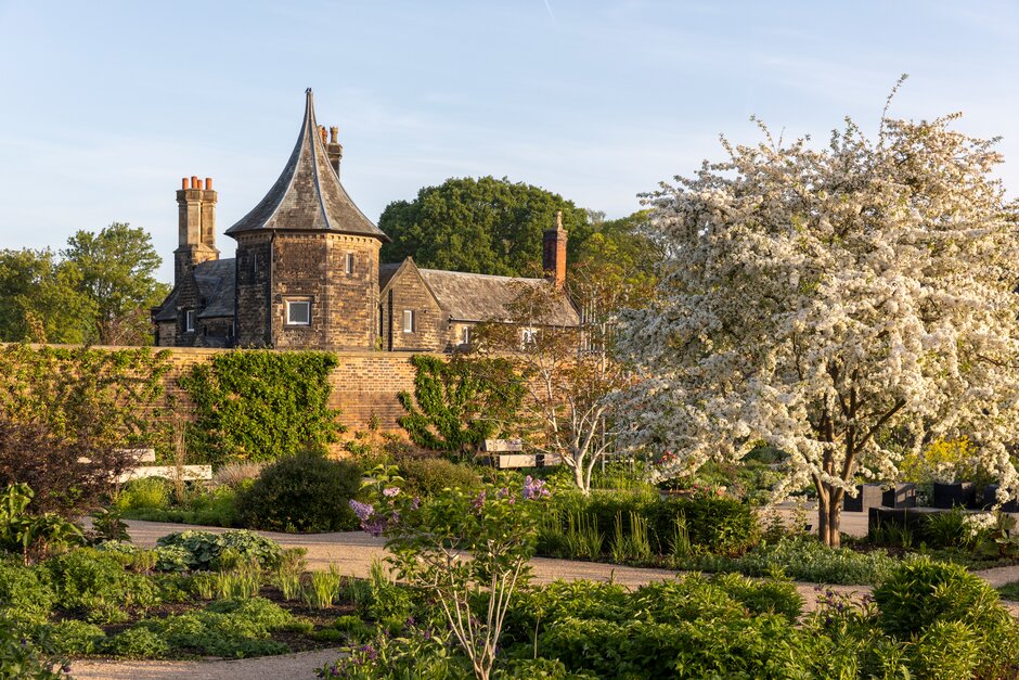 Historic stone building overlooking landscaped gardens in spring, with a flowering tree, paths and benches in the foreground.