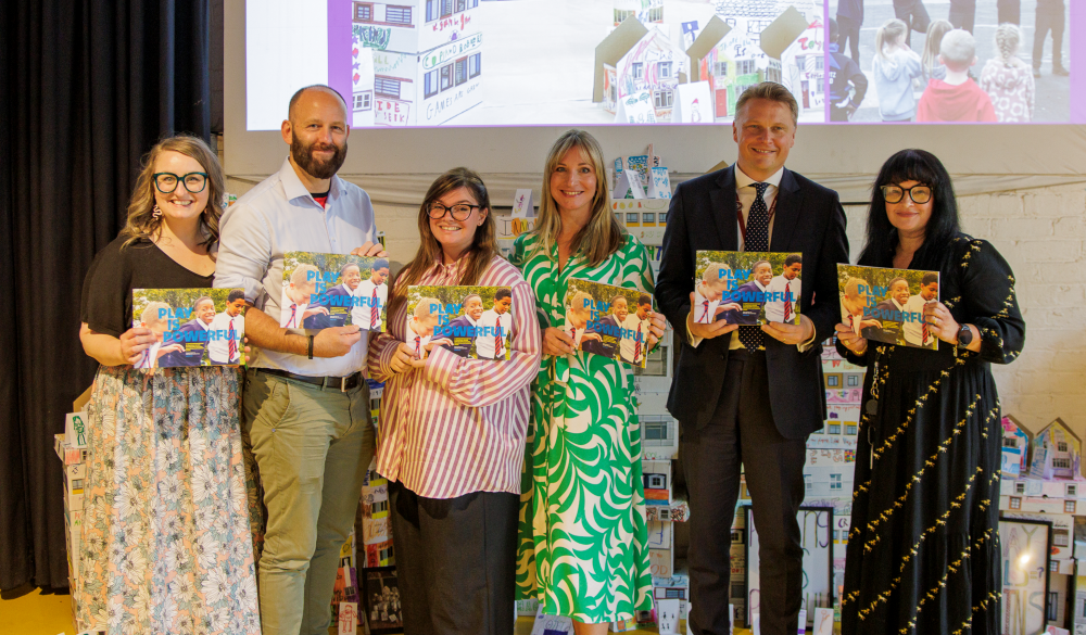 Salford Cultural Education Partnership partners stands in a line, smiling and holding up booklets with the cover text "PLAY IS POWERFUL" featuring a photo of schoolchildren. They are standing in front of a wall decorated with miniature paper houses and a projected screen showing images of children's artwork and activities.