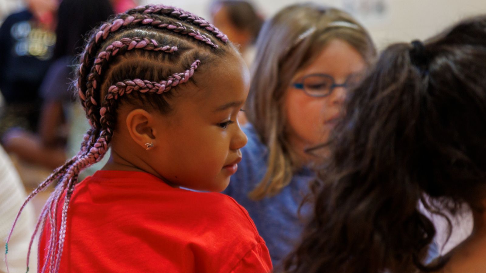 Child with pink corn rows and red top with two other children.