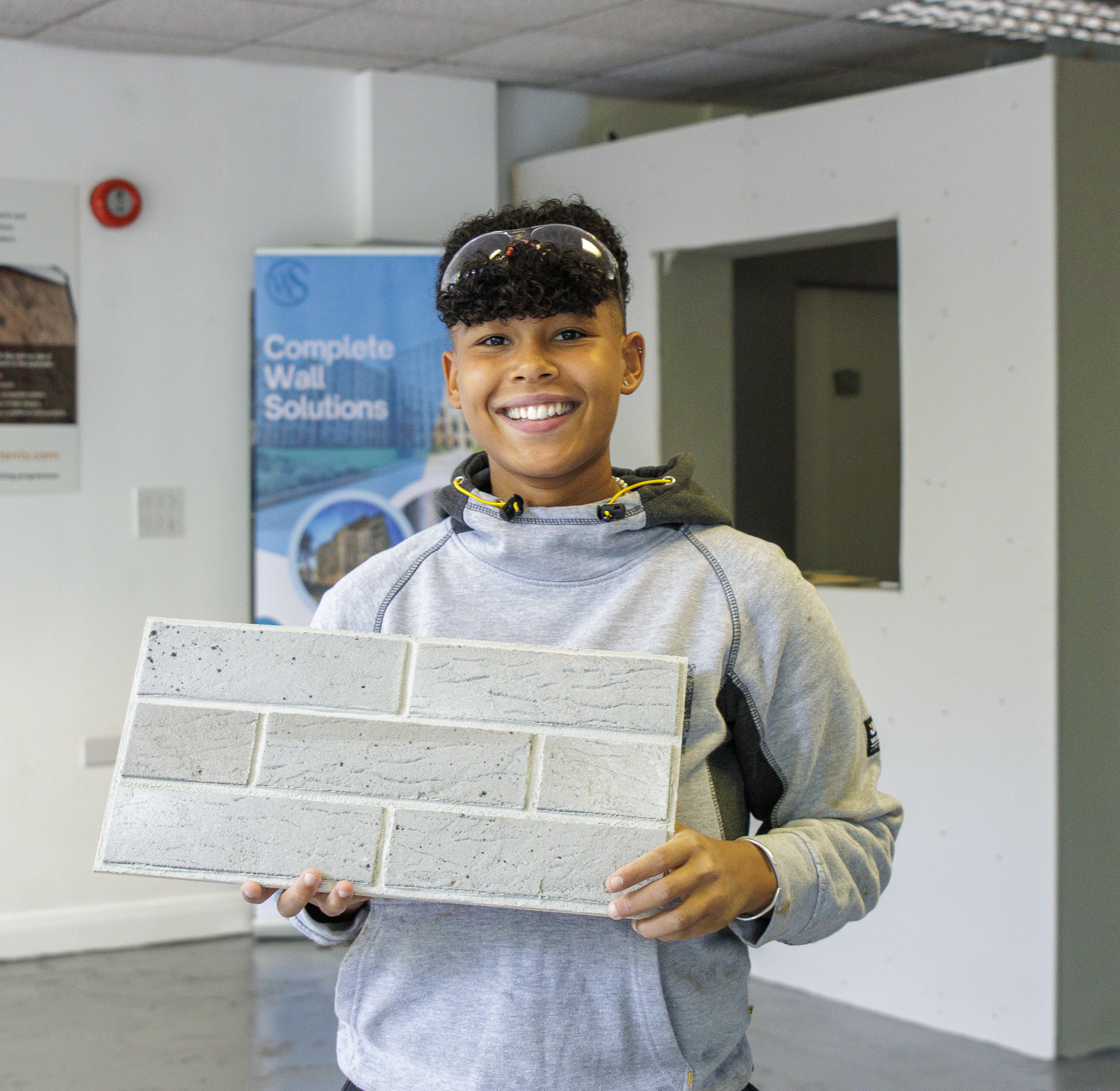 A young lady smiles and shows her brickwork sample. She is wearing safety goggles on her head and a grey hoodie. In the background a plastering bay and a blue banner displaying the text 'Complete Wall Solutions' can be seen.