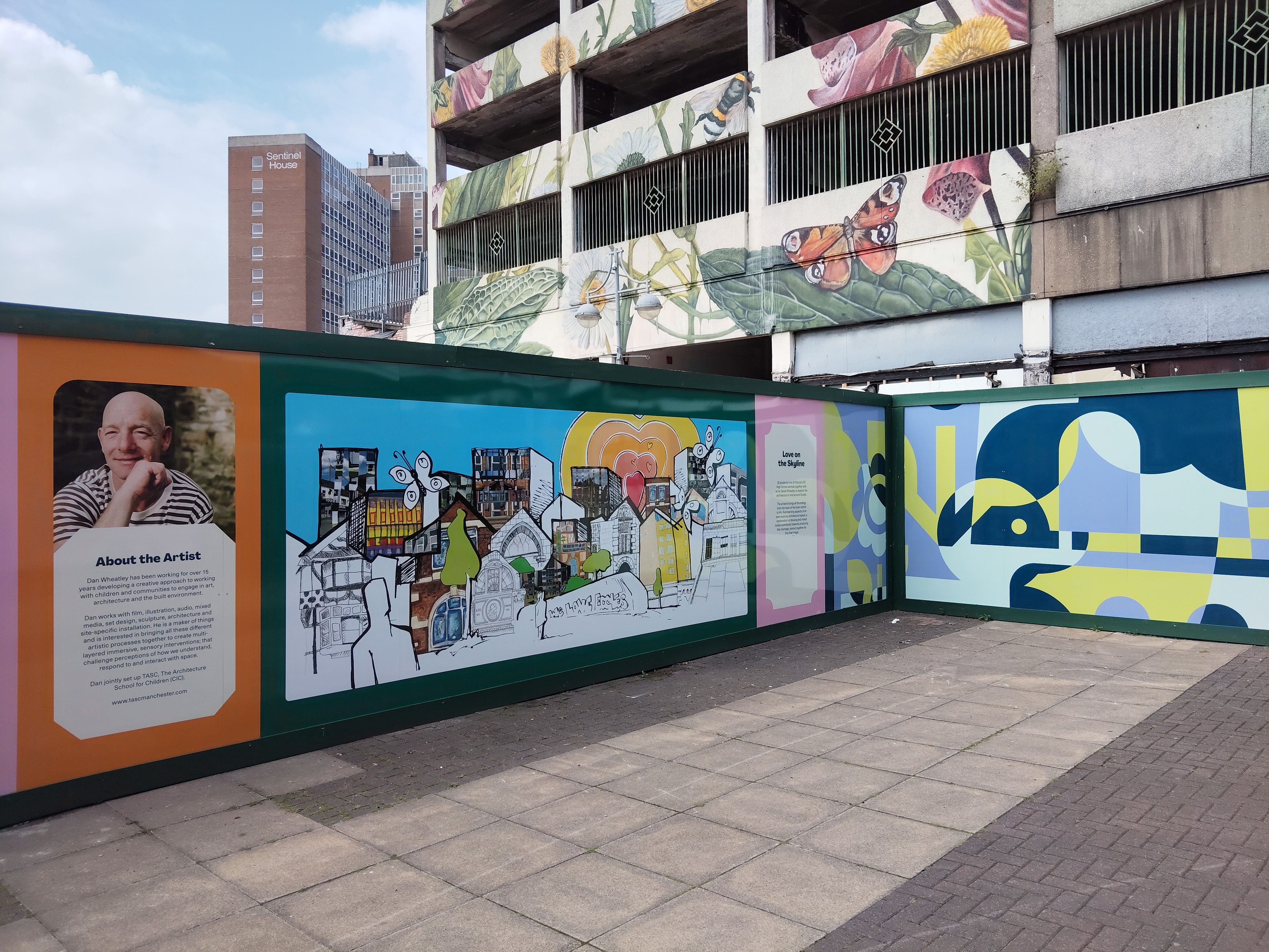A section of an outdoor mural on a building wall featuring colorful artwork and text panels. The mural includes an 'About the Artist' panel on the left, a vibrant cityscape illustration in the center, and abstract geometric patterns on the right.