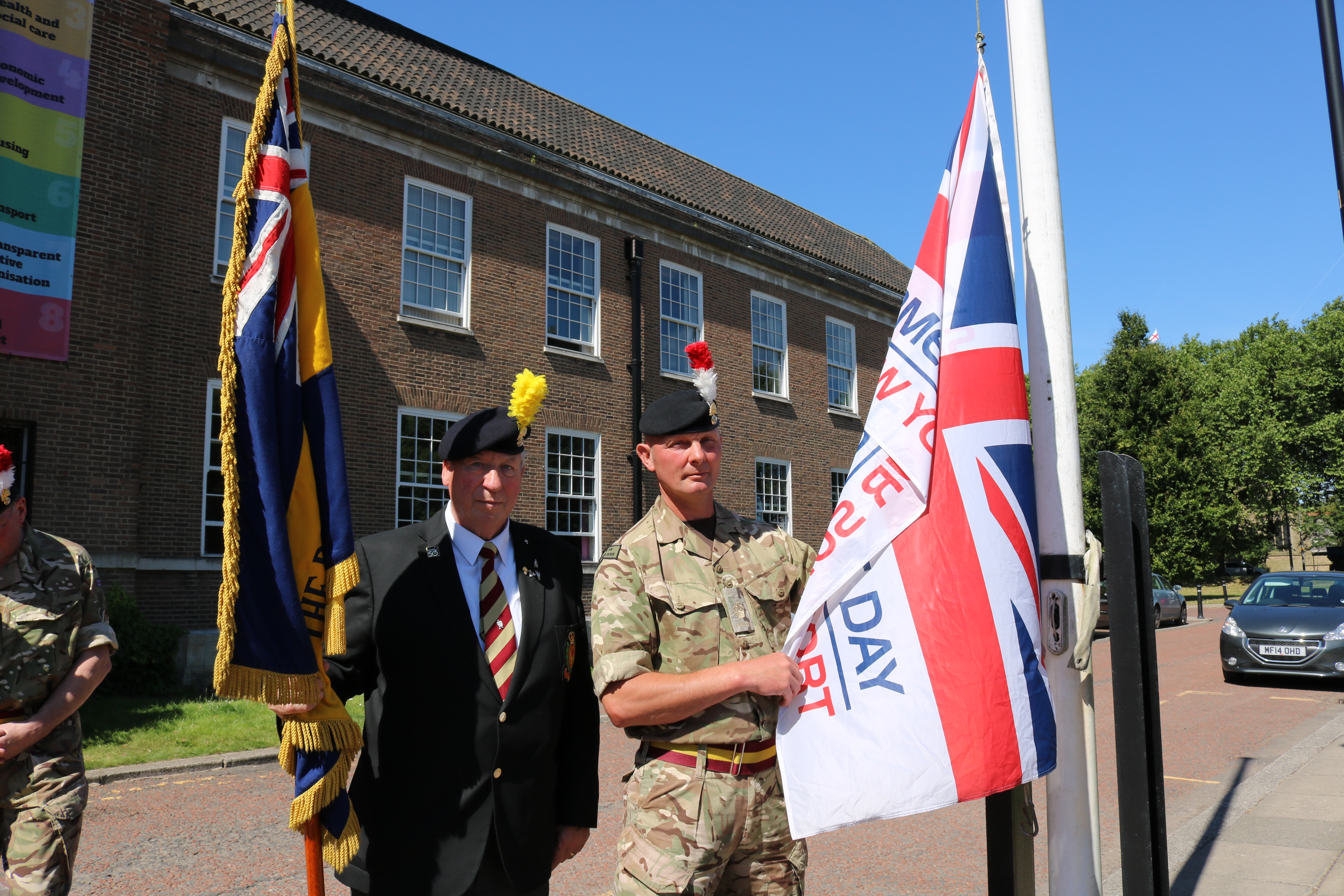 Armed Forces personnel stood in uniform holding Armed Forces flag outside Salford Civic Centre on a sunny day with blue skies in the background