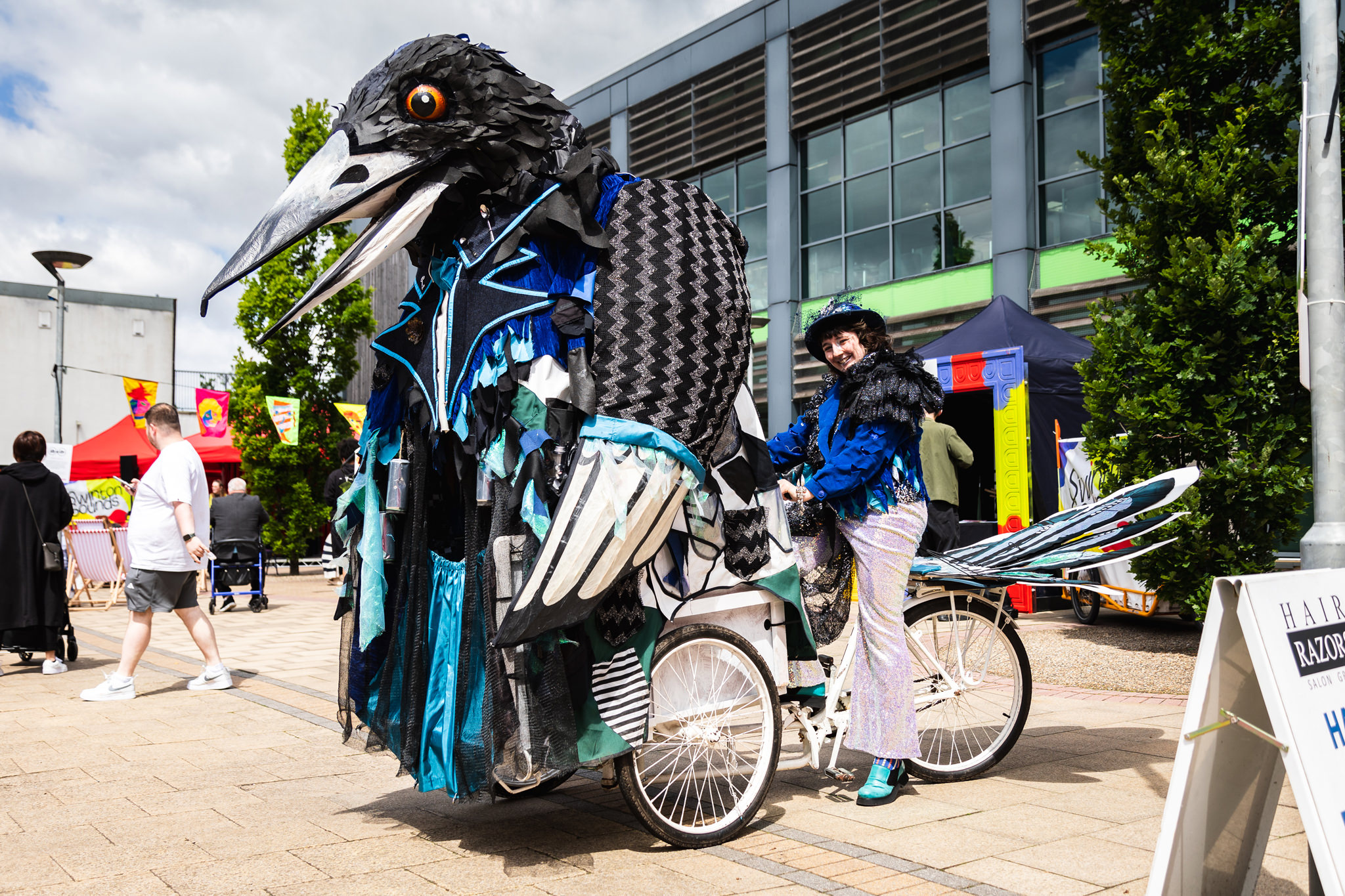 A person rides a bicycle with a giant bird sculpture perched on the front, in Swinton Square. Various entertainment in the background, with people sat in deckchairs.