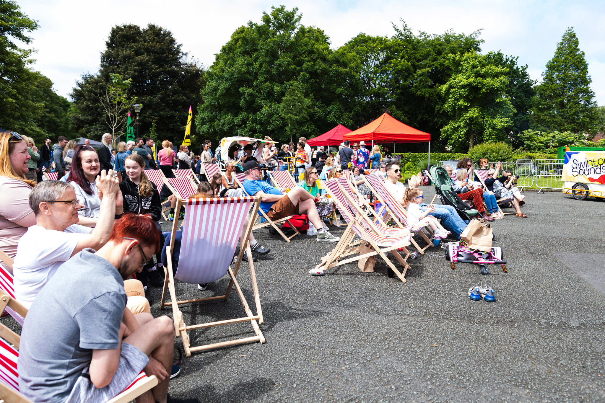 A group of people sitting on deckchairs outdoors, surrounded by trees under a clear sky, enjoying Swinton Sounds 2024 in Victoria Park, Swinton.