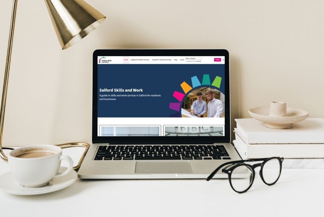 A laptop screen displaying the Salford Skills and Work website. The laptop is sat on a desk next to a gold lamp, a coffee cup, a pair of glasses, a stack of books and a candle stick holder.