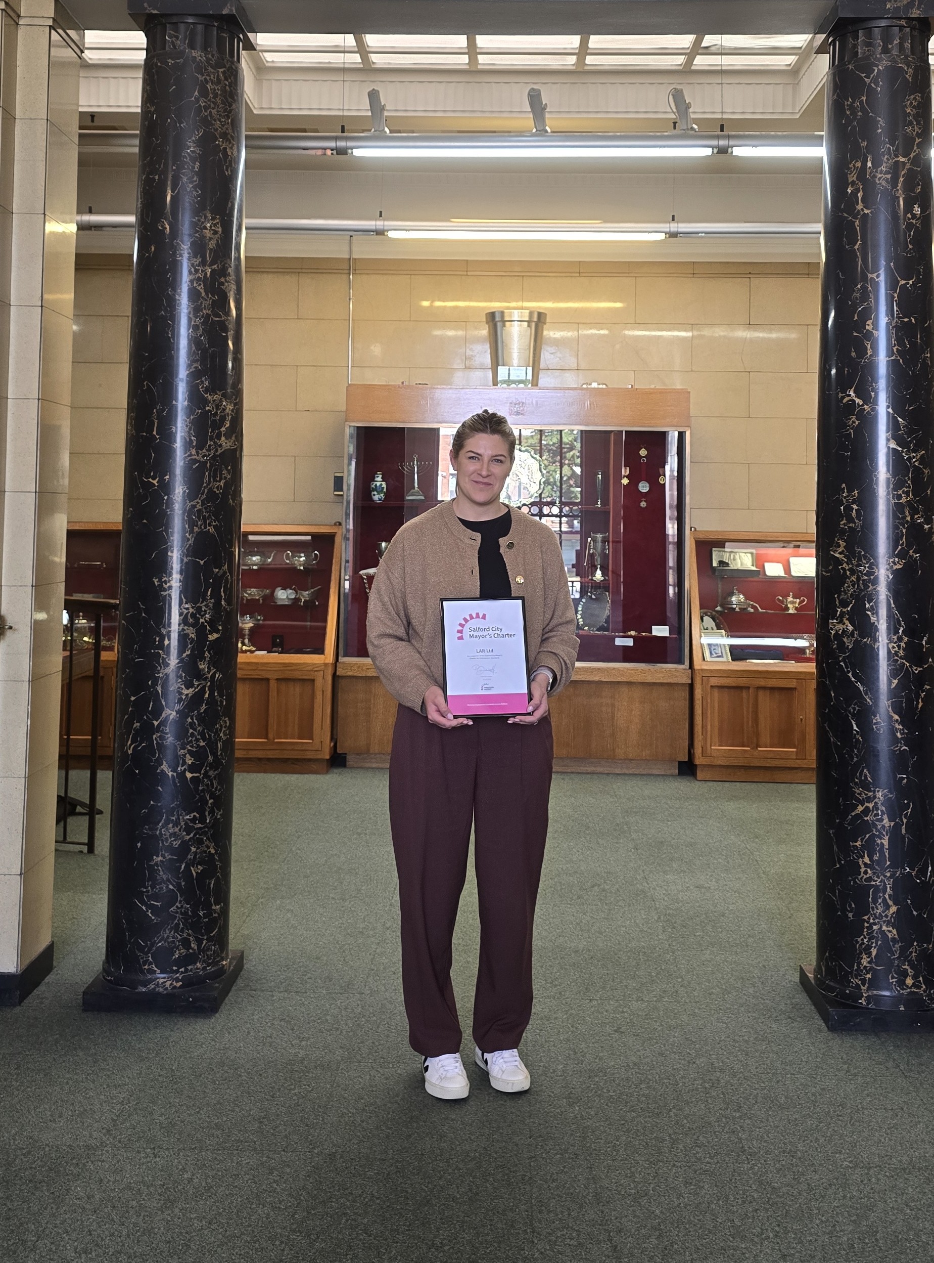 A woman stands smiling holding a framed certificate. She is wearing a brown cardigan and black trousers. In the background are some wooden cabinets containing trophies.