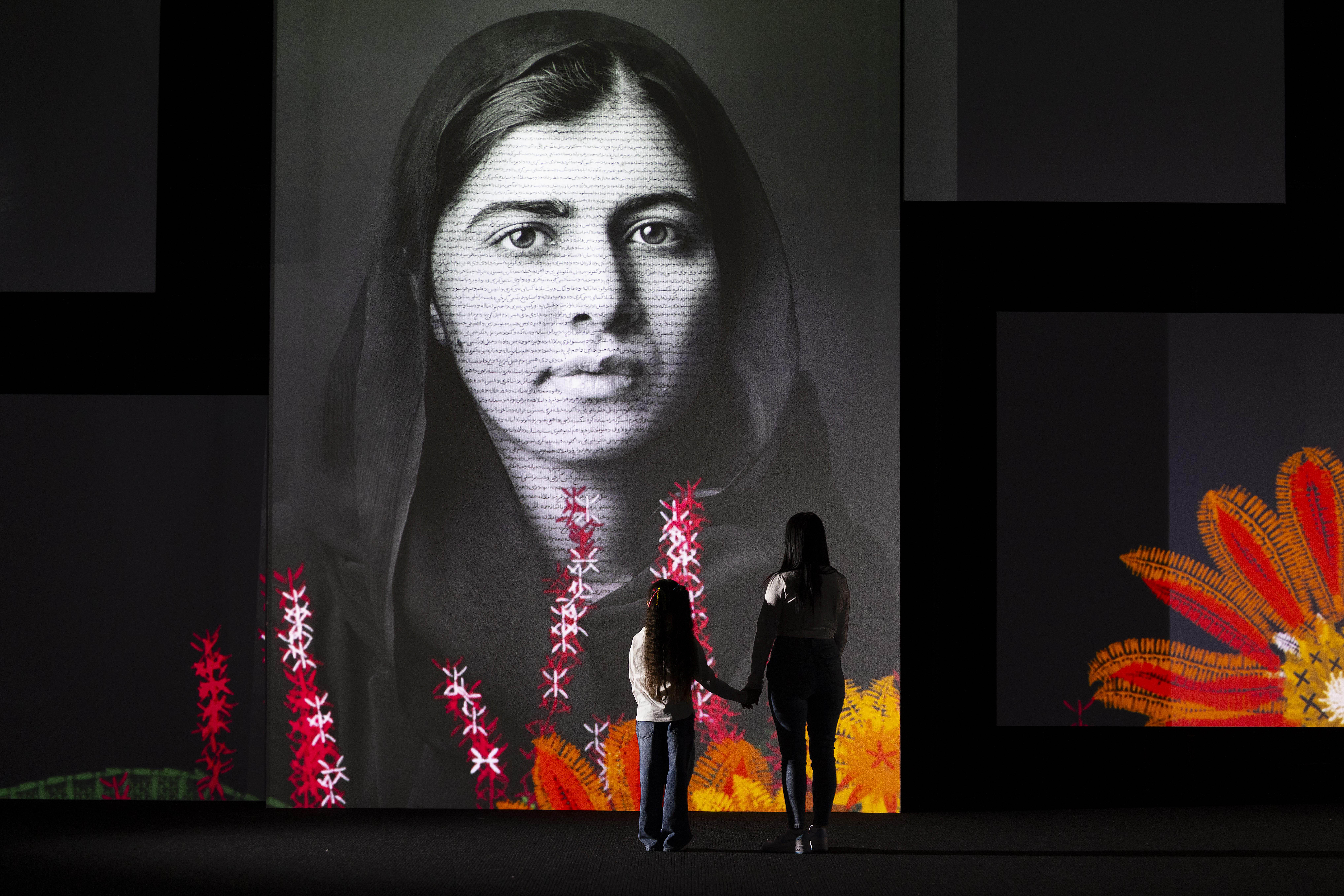 Large scale black and white portrait of Malala Yousafzai on screen towering over an adult and child in foreground. There are red, orange and white digitally illustrated flowers blooming from the ground.