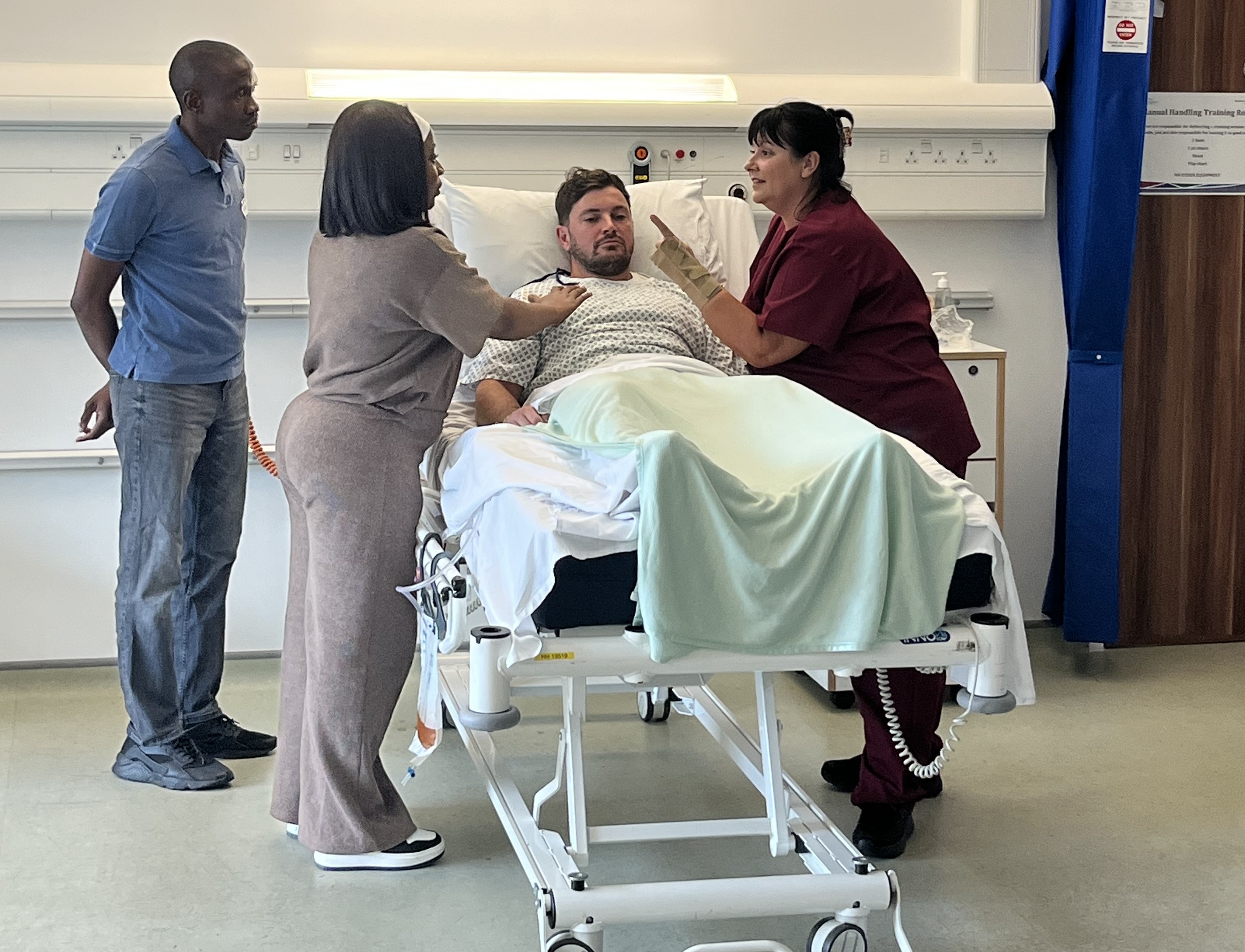 A man lies in a hospital bed with a man and two women stood over him having a conversation