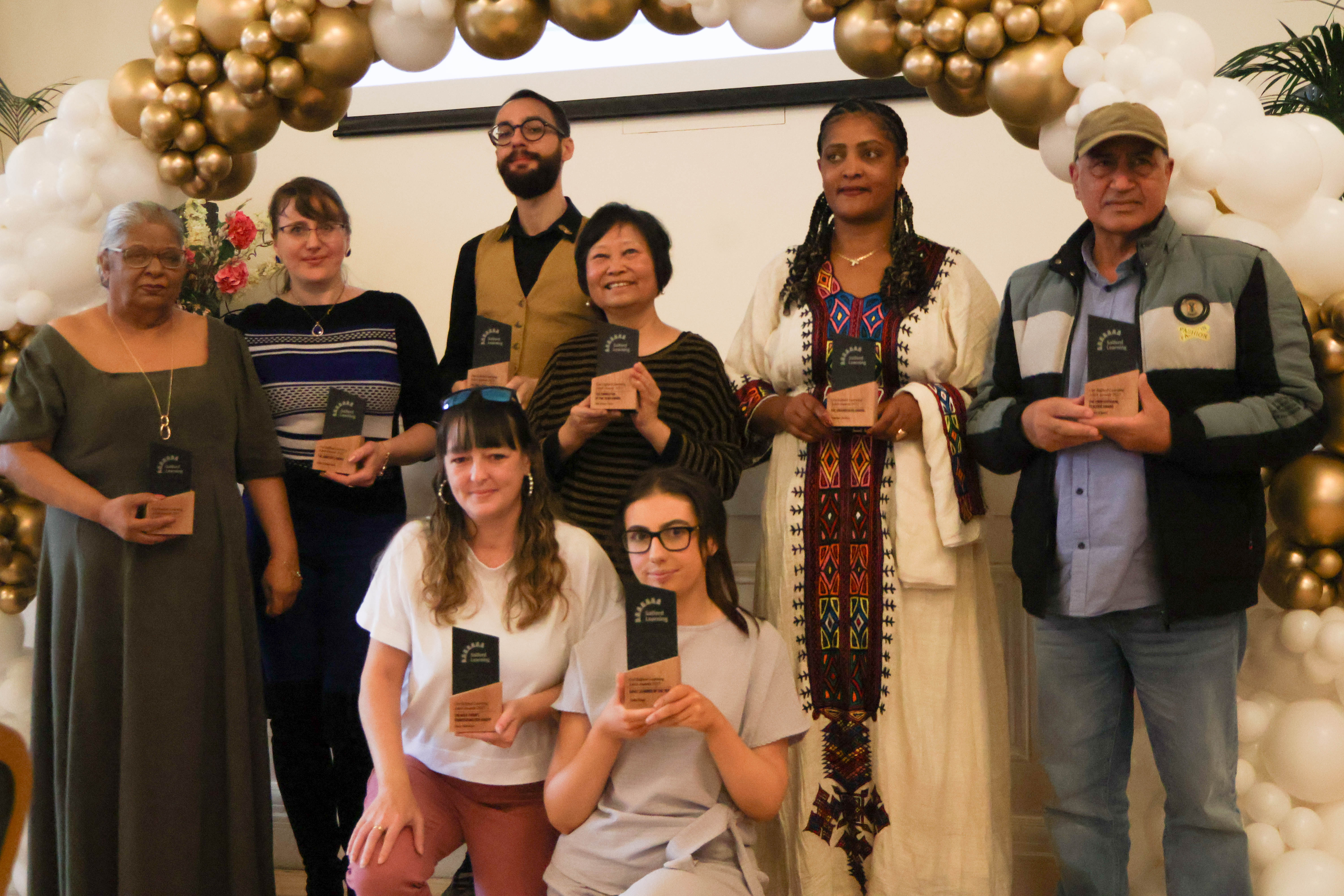 A group of smiling people stood under a white and gold balloon arch