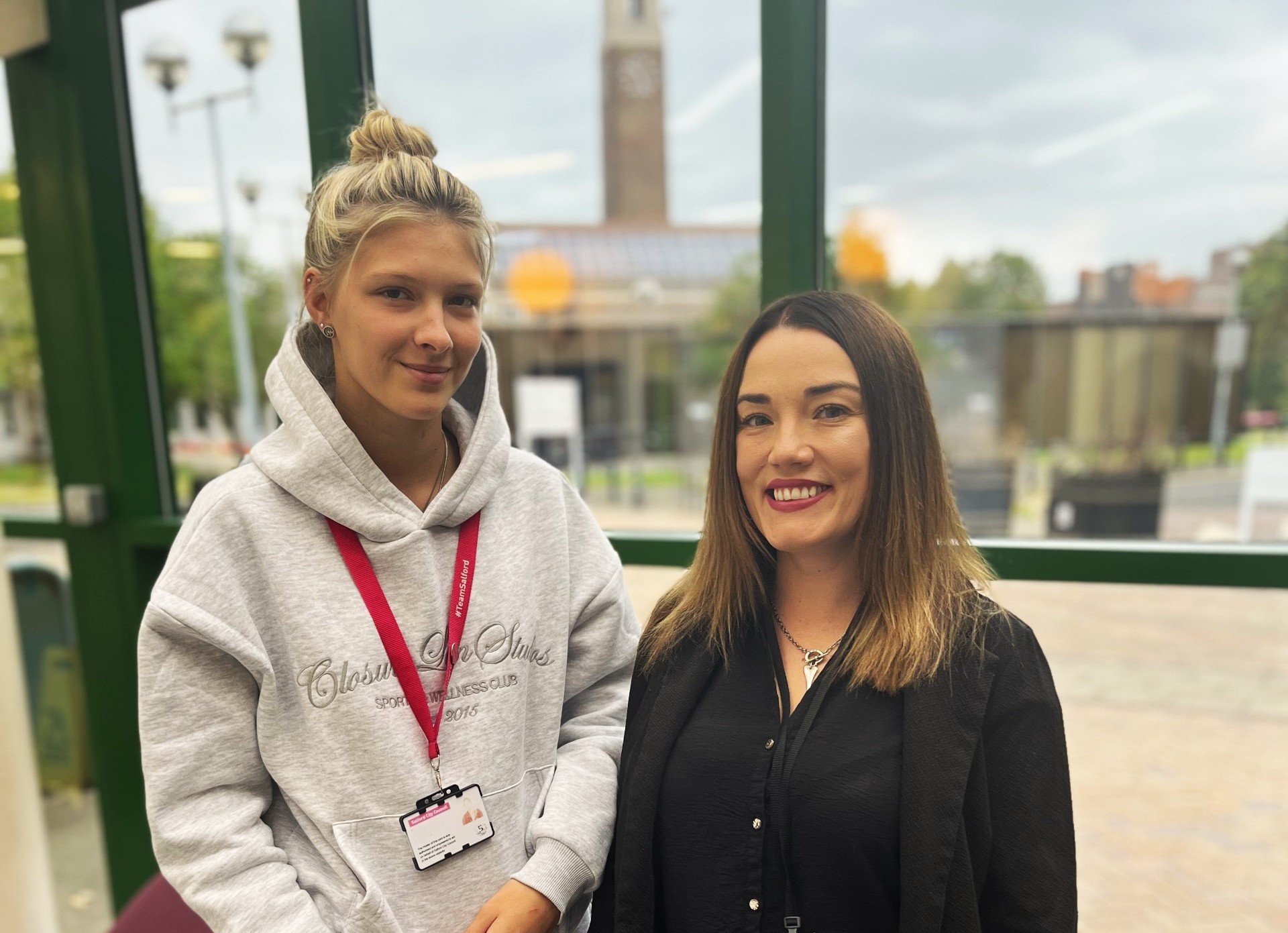 Two women stand in front of a window smiling. Behind them, a clock tower can be seen out of focus.