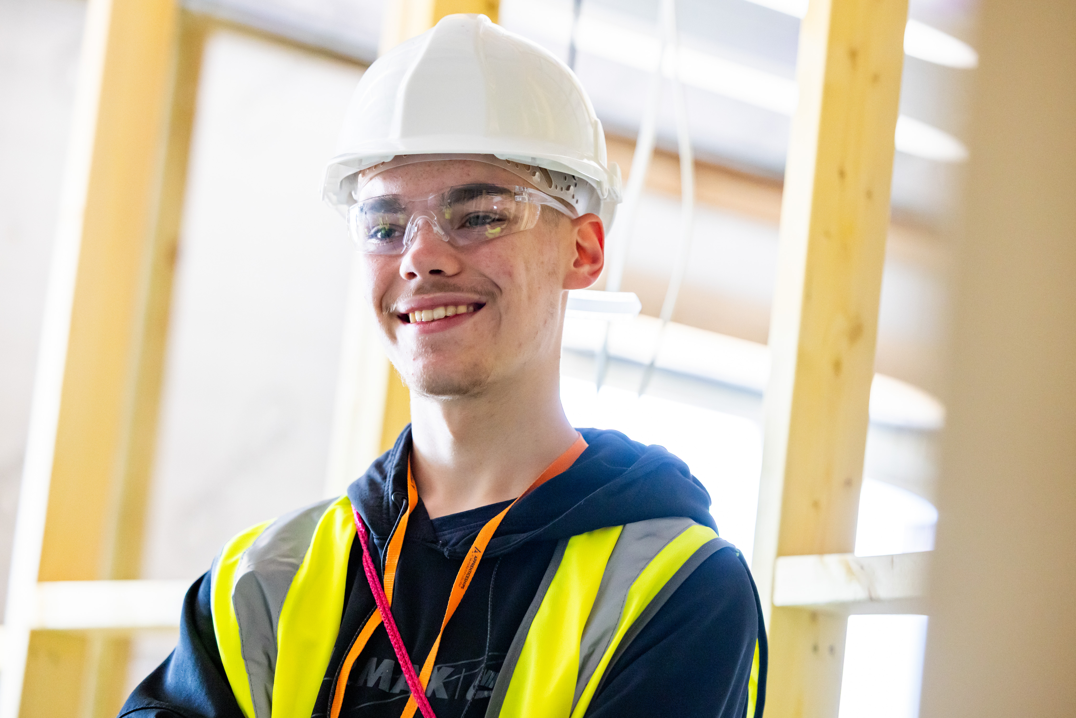 A young man wearing a hard hat, protective glasses, a navy blue hoody and a hi-vis vest smiles whilst being stood on a housing construction site