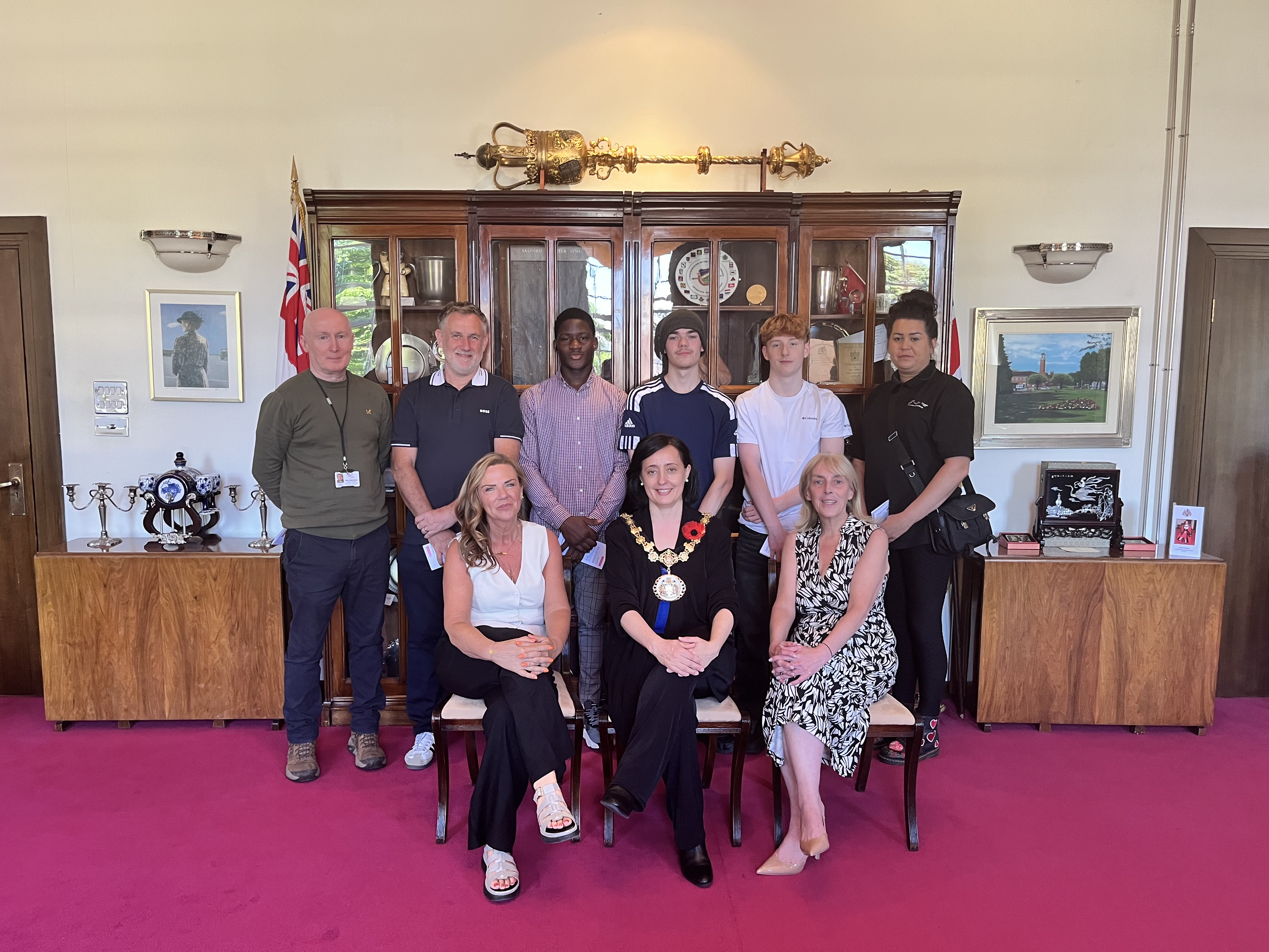 A group of smiling people stood and sat in the Ceremonial Mayor's office in front of a large brown cabinet. On top of the cabinet lies an ornamental gold mace.