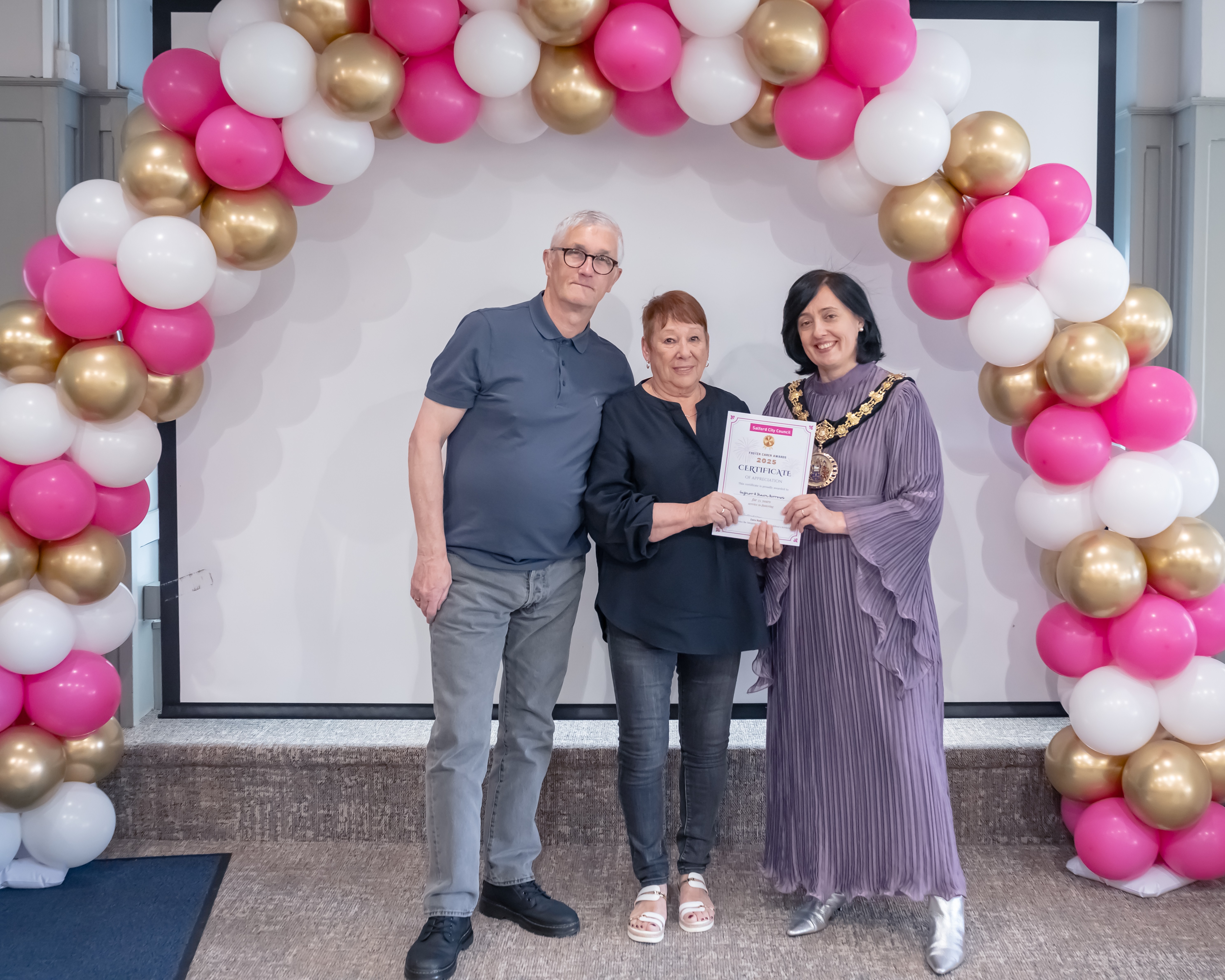 A man and woman stood with the Cermemonial Mayor of Salford Councillor Tanya Burch. A balloon arch is behind them made up of white, gold and magenta balloons.