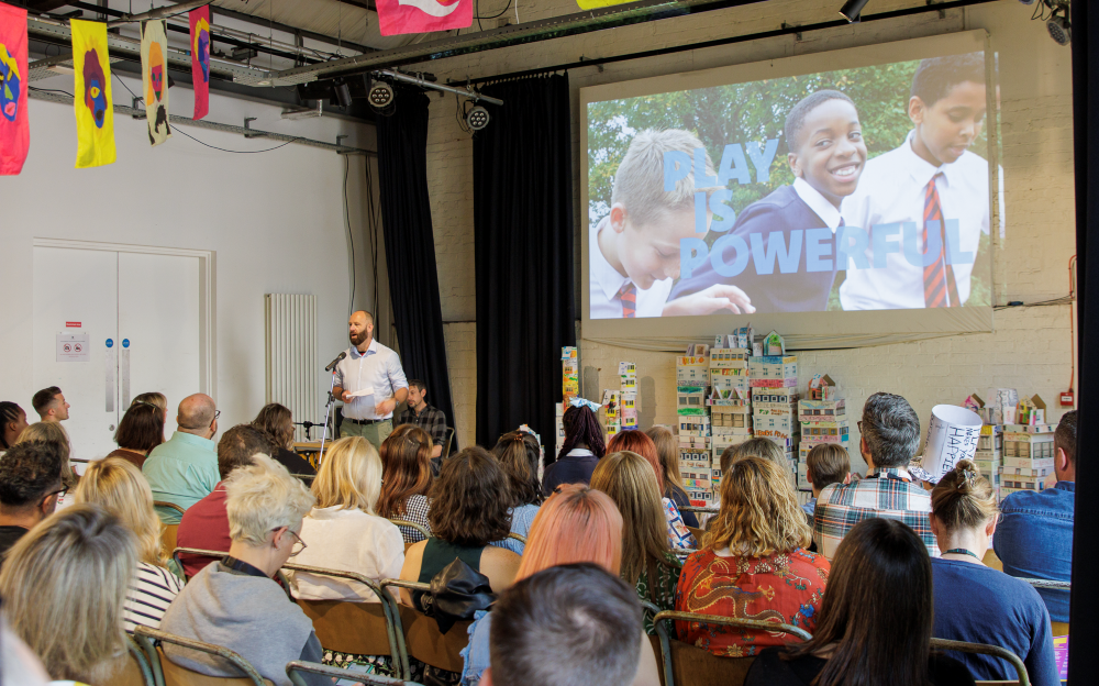 Paul Dennett, Salford City Mayor, stands at a microphone addressing an audience seated in a room with colourful banners hanging from the ceiling. Behind him, a large screen displays an image of three school-aged boys in uniforms with the words "PLAY IS POWERFUL" overlaid. The wall under the screen is decorated with numerous small model houses made from cardboard or paper. The photo was taken at the Salford Cultural Education Partnership strategy launch event.