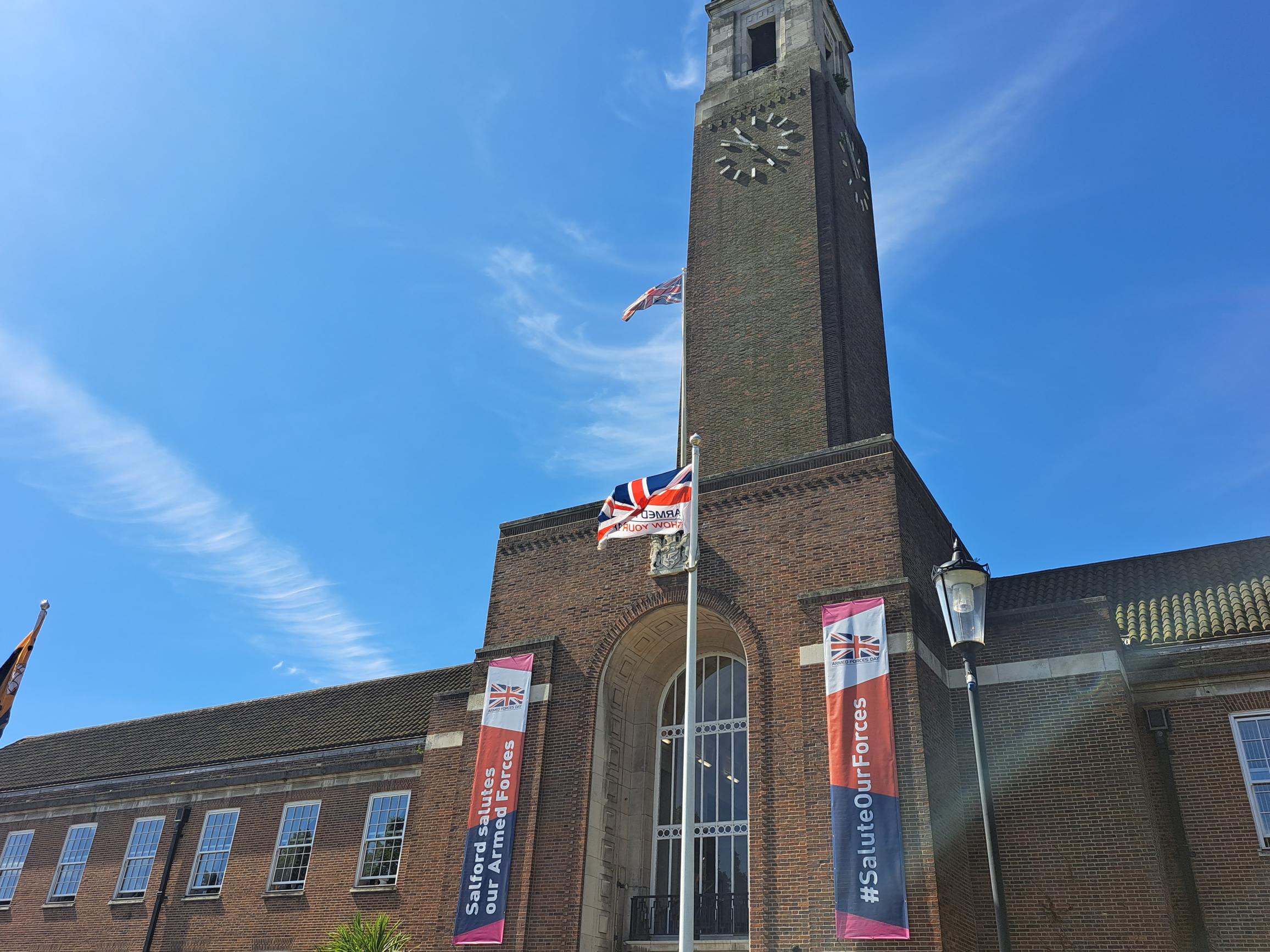 Image of Salford Civic Centre featuring the clock tower decorated with red, white and blue Armed Forces town hall banners and the flag pole with the Armed Forces Day flag flying high in the wind. Bright blue sunny skies also feature in the background