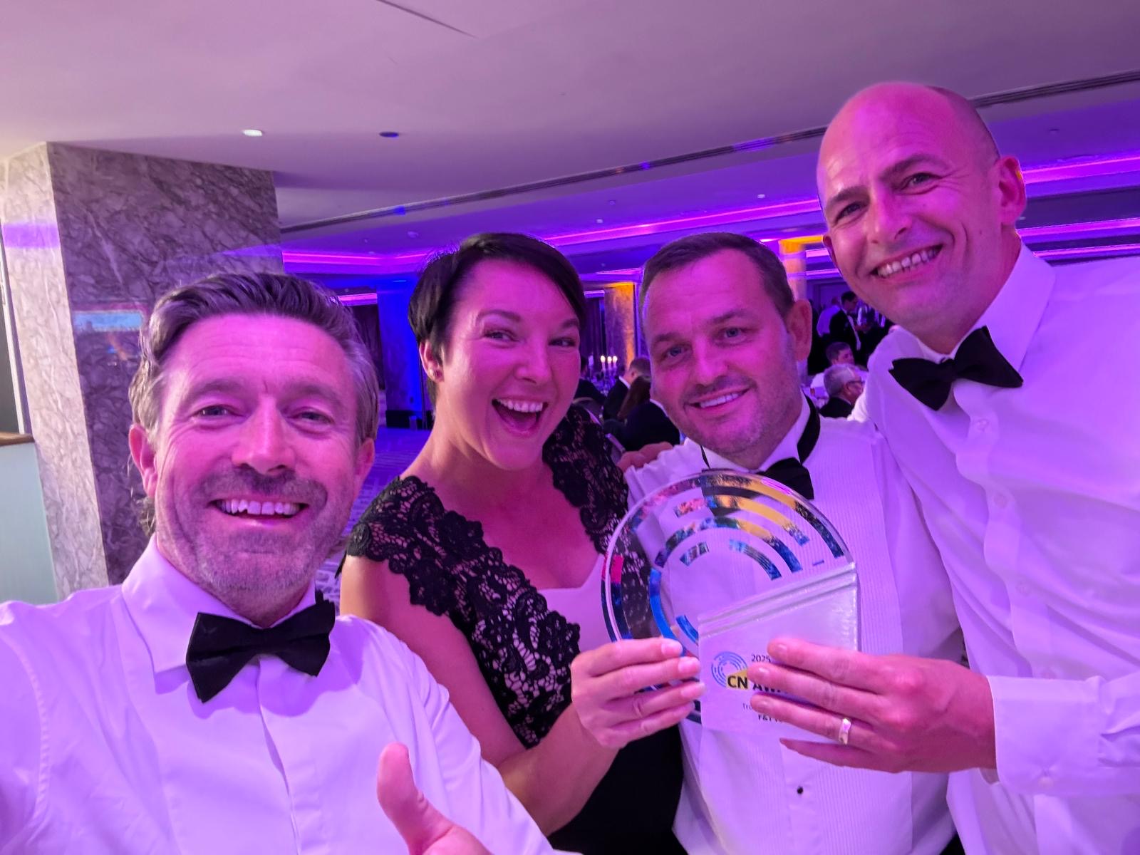 Three men and a woman stood smiling and posing with a glass trophy in purple lighting. The three men are wearing white shirts and black bow ties. The woman is wearing a black dress.