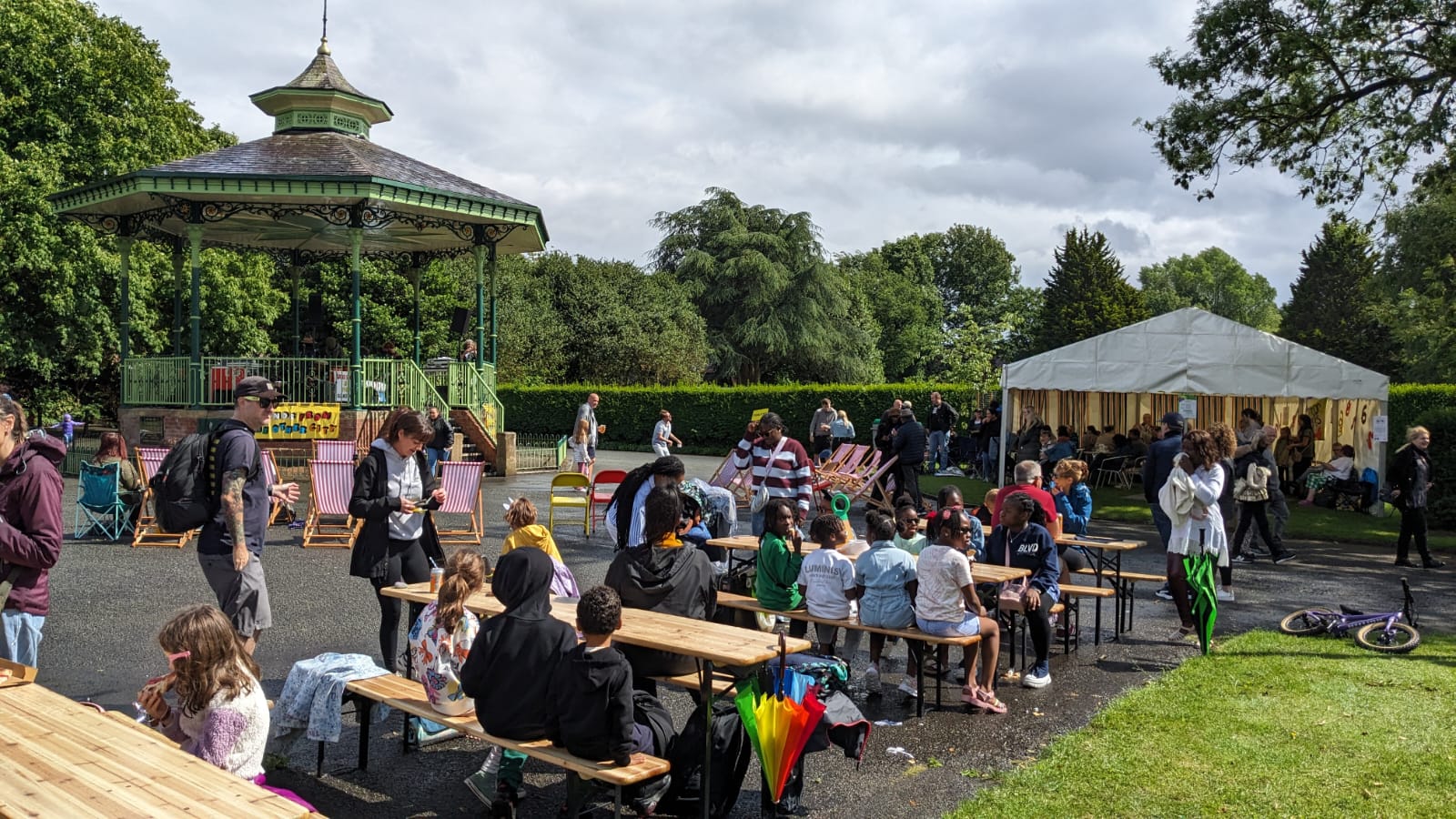 A lively outdoor community event at Victoria Park with families and children sitting at wooden picnic tables. A green bandstand in the background hosts performers, and a white marquee tent is set up on the right. People are walking, chatting, and enjoying the festivities under a cloudy but bright sky.
