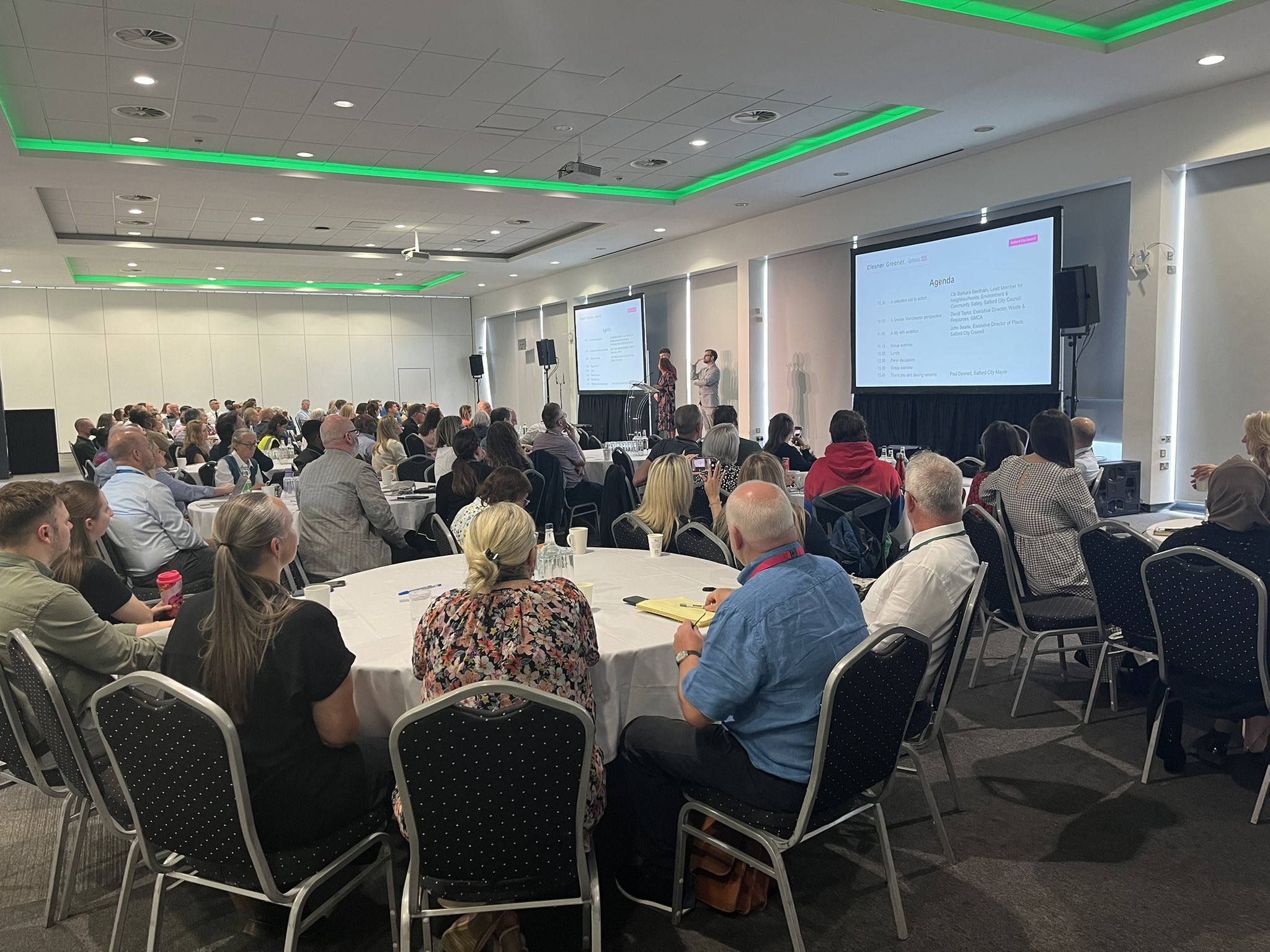 A room full of attendees sat a large round tables faced towards two large screen displaying the agenda for the Cleaner Greener Conference.