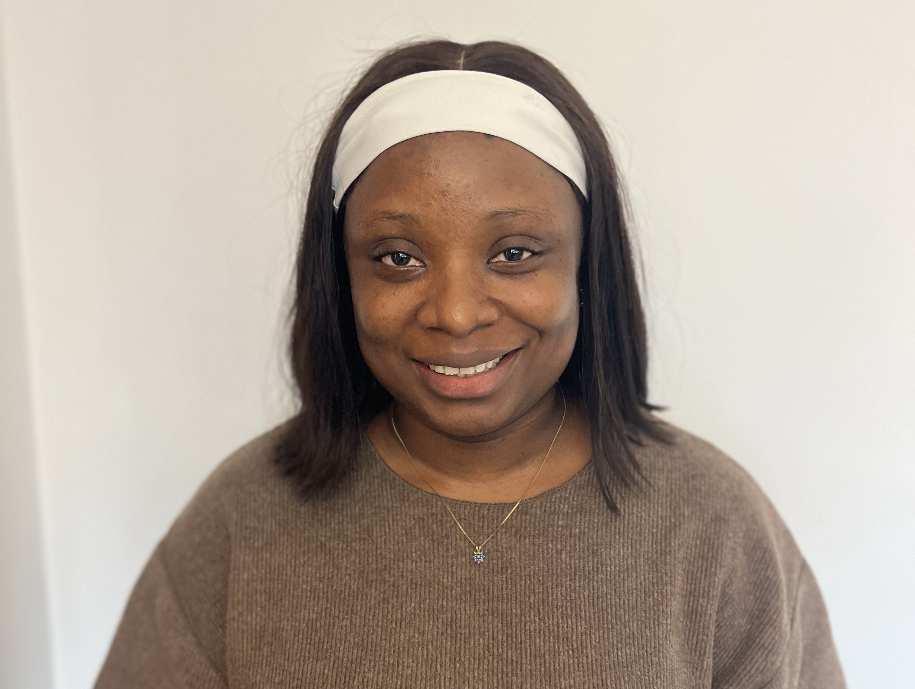 A woman stands smiling against a white wall. She is wearing a white headband and a brown knitted jumper.